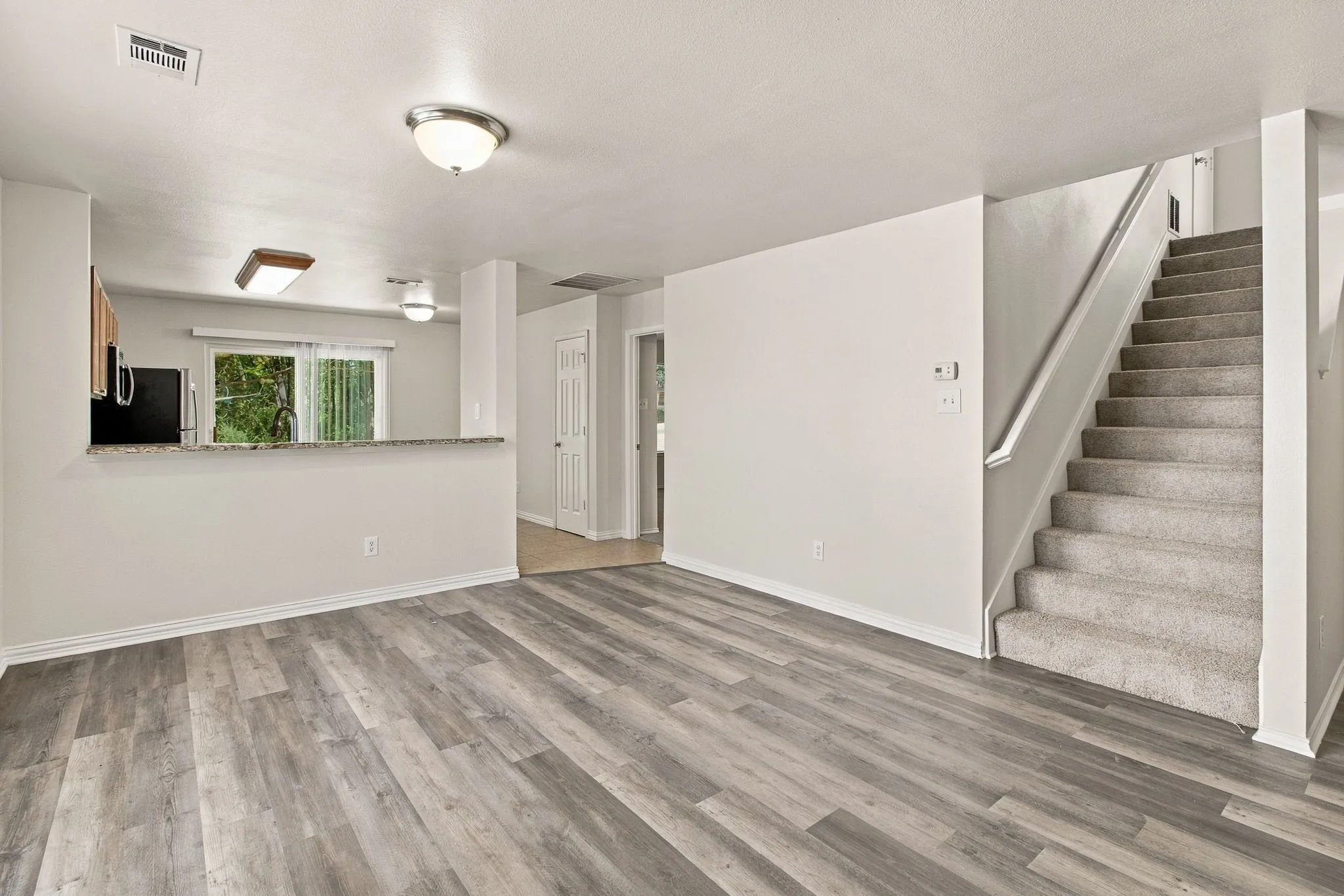Wood-finish flooring throughout the main area, a carpeted staircase with a handrail, and a kitchen pass-through featuring a granite-style countertop