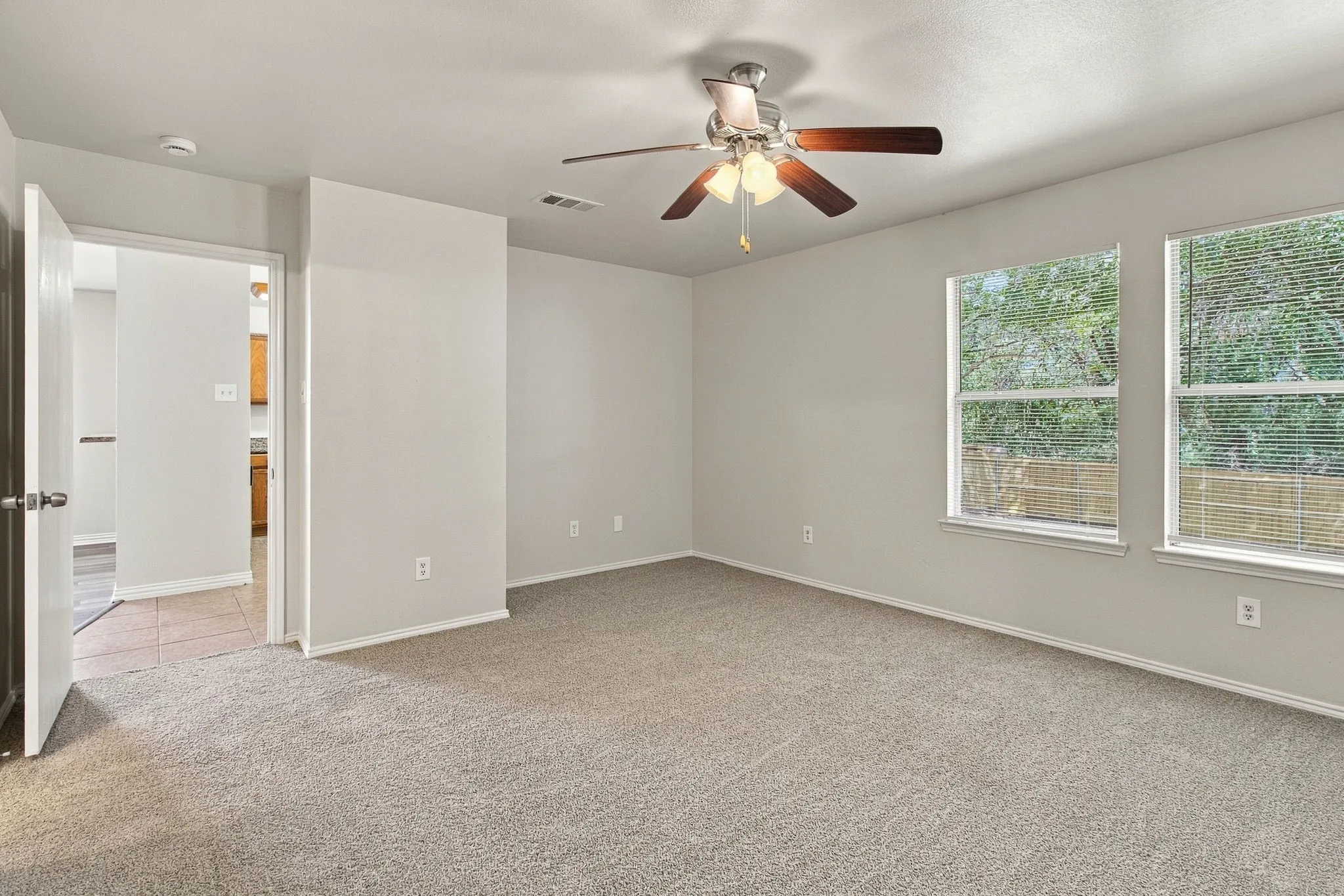 Expansive carpeted room featuring a ceiling fan with light fixture, two windows with blinds, and white trim