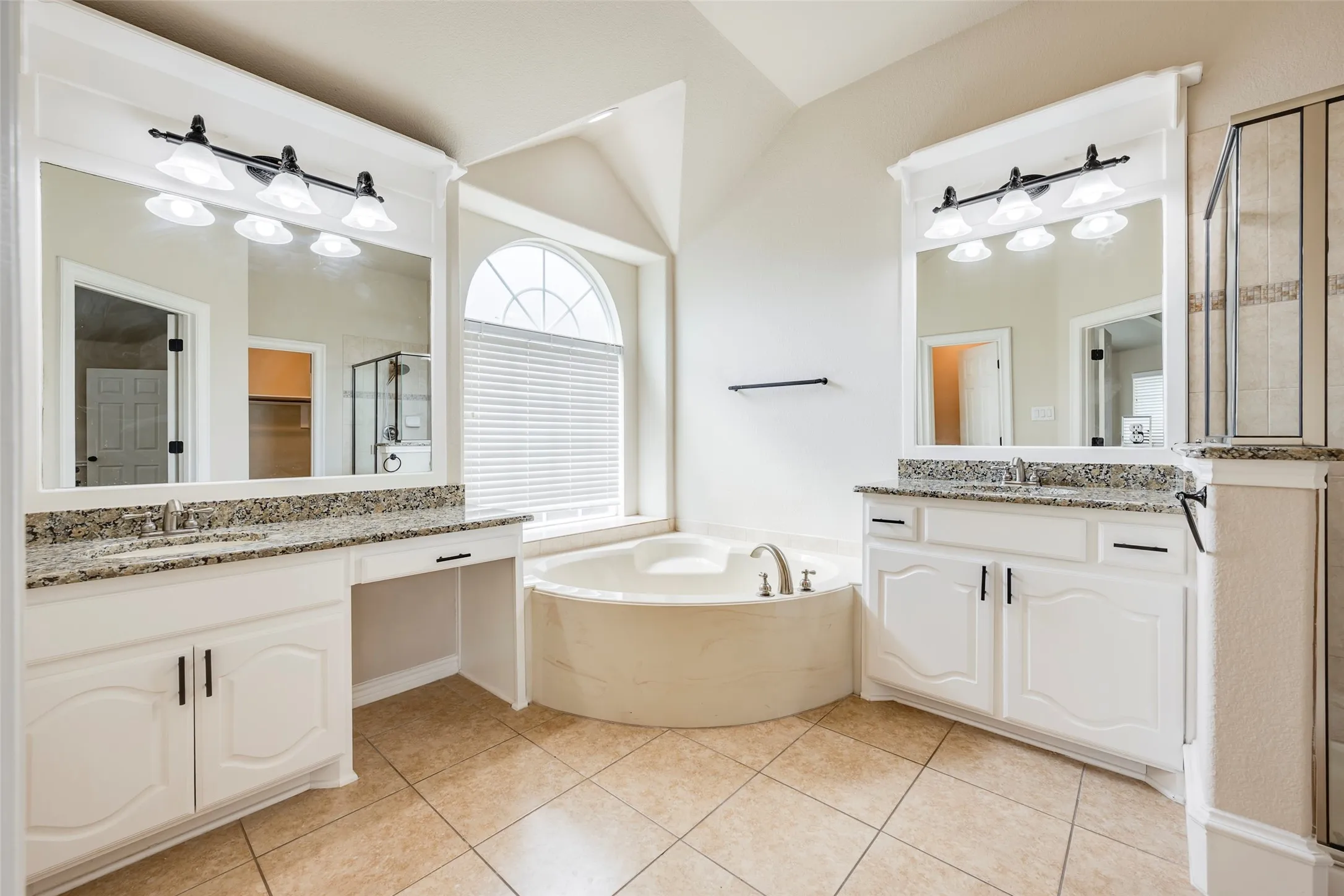 Dual vanities with granite countertops, an arched window, a built-in soaking tub, and tile flooring