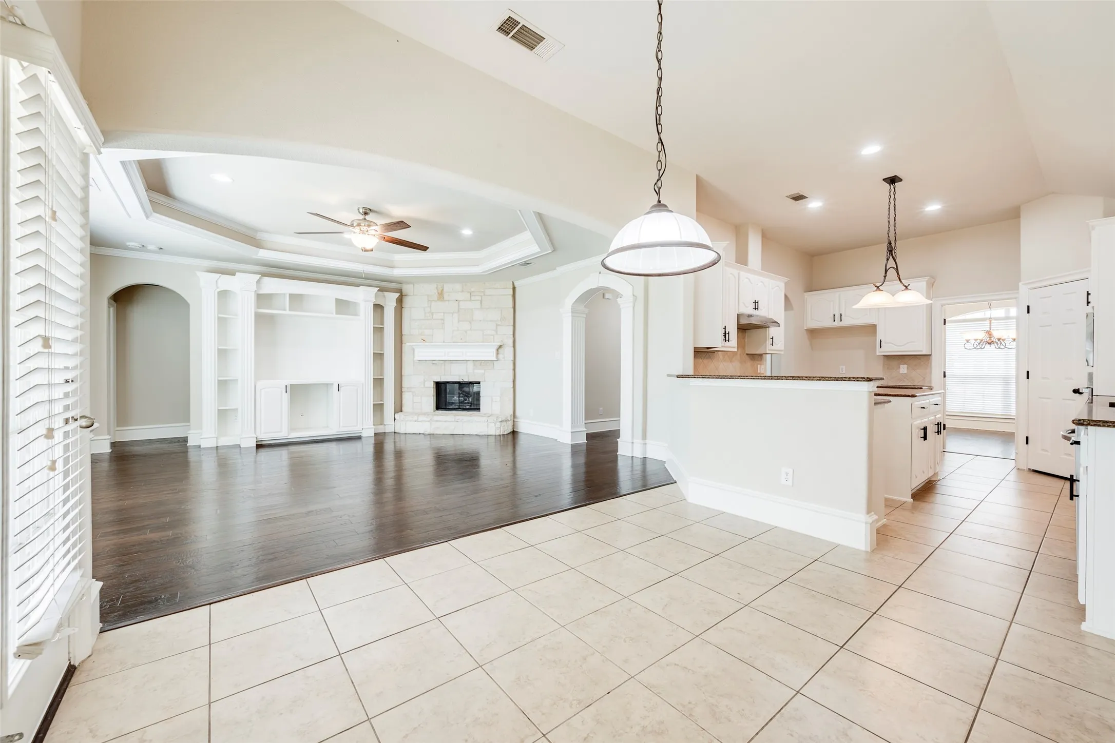 Vaulted ceiling with a fan, stone-surround fireplace, built-in shelving, wood-finish flooring, and a kitchen with pendant lighting