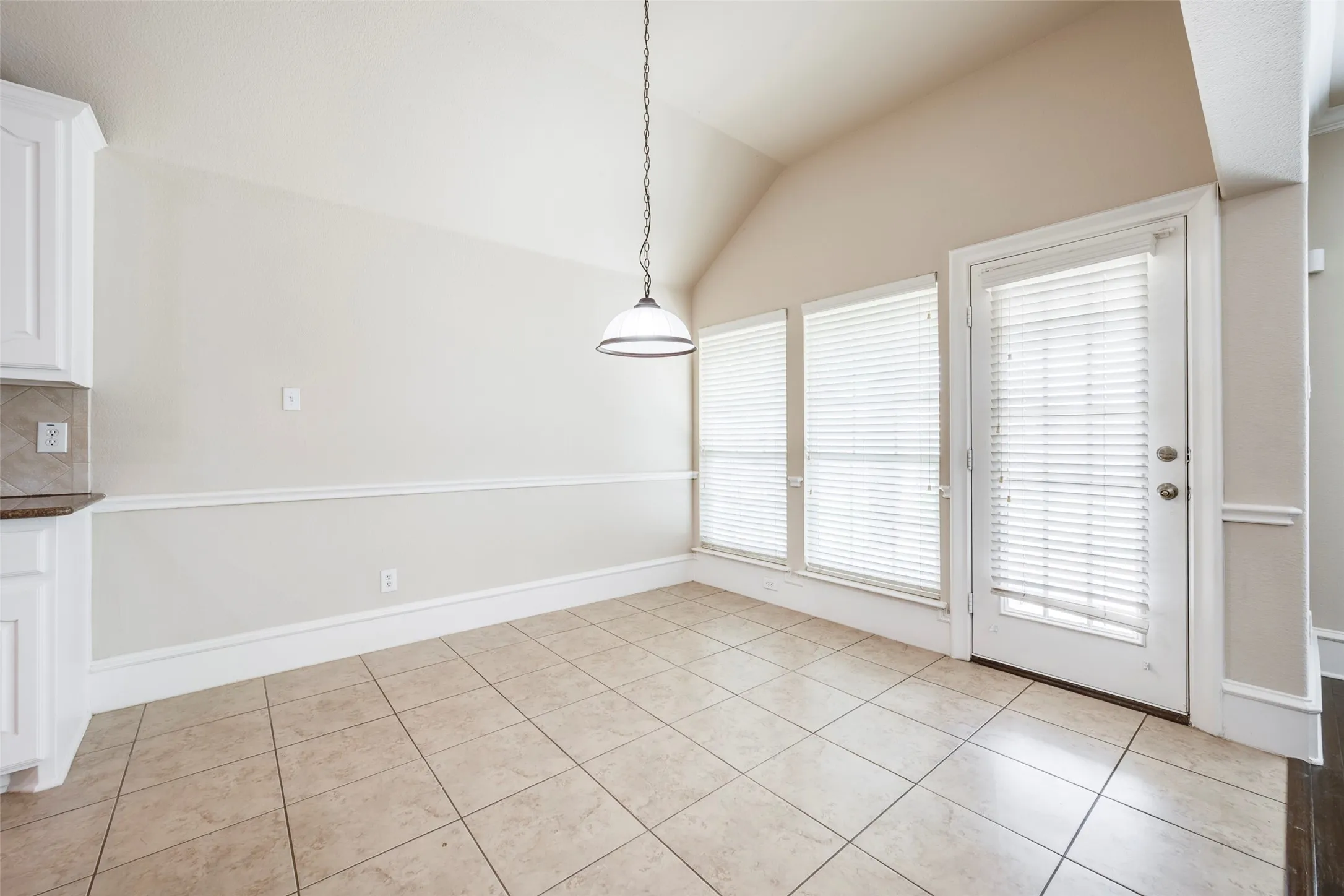 Vaulted ceiling, pendant light fixture, three windows, glass-paneled door, and tile flooring