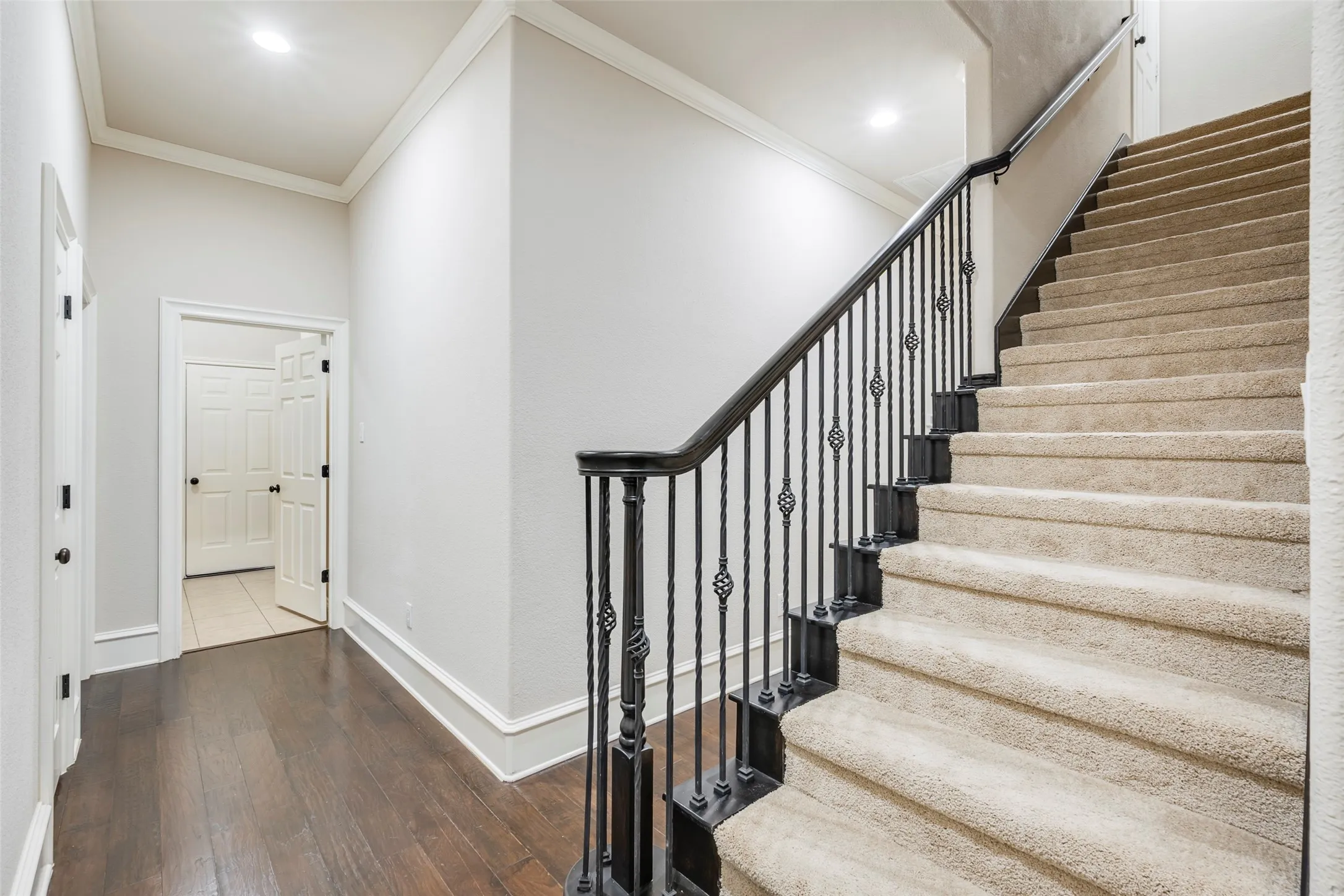 Carpeted staircase with wrought iron balusters, dark wood handrail, recessed lighting, crown molding, and wood-finish flooring
