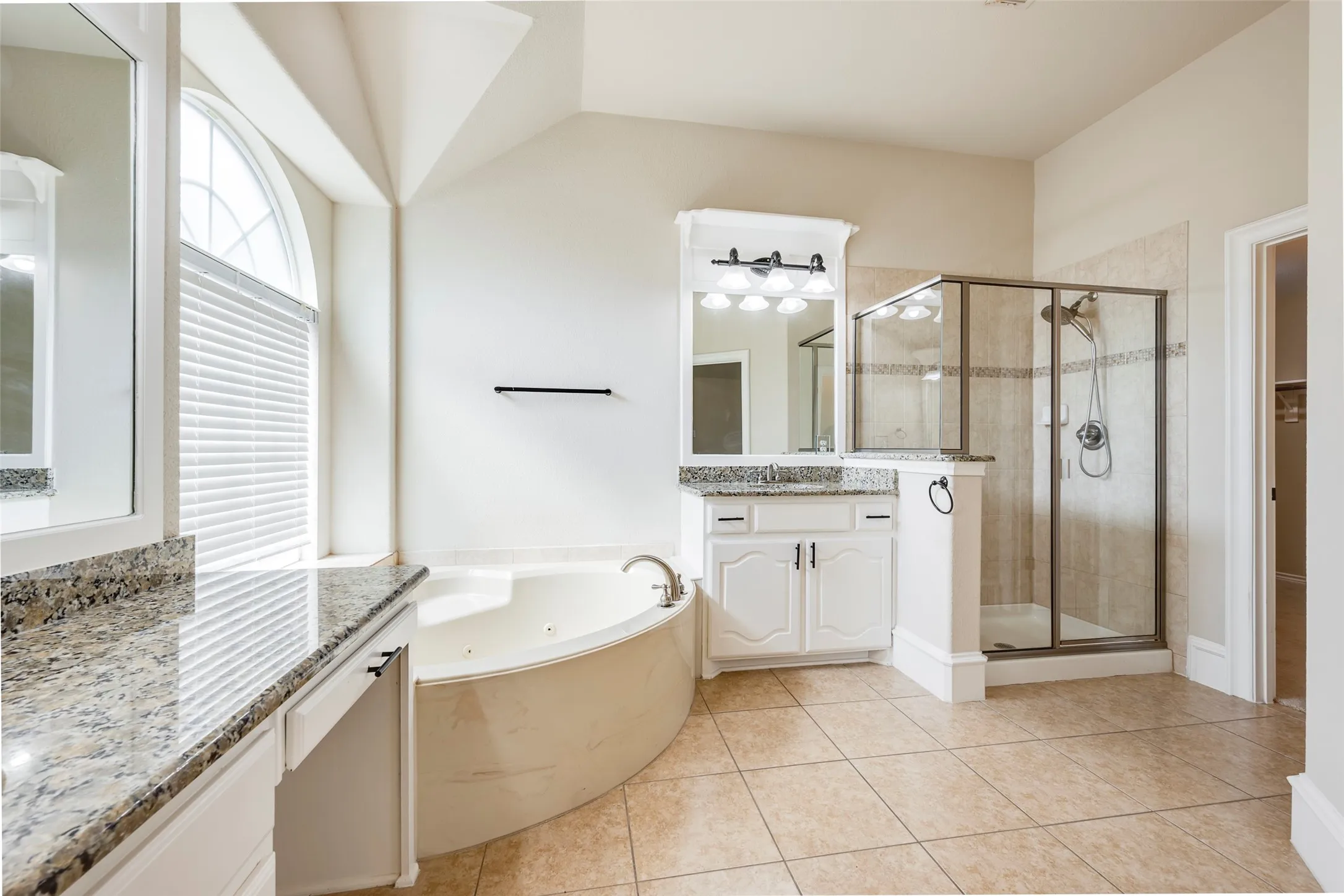 Arched window with blinds, built-in soaking tub, glass-enclosed shower, dual vanities with stone countertops, and tile flooring