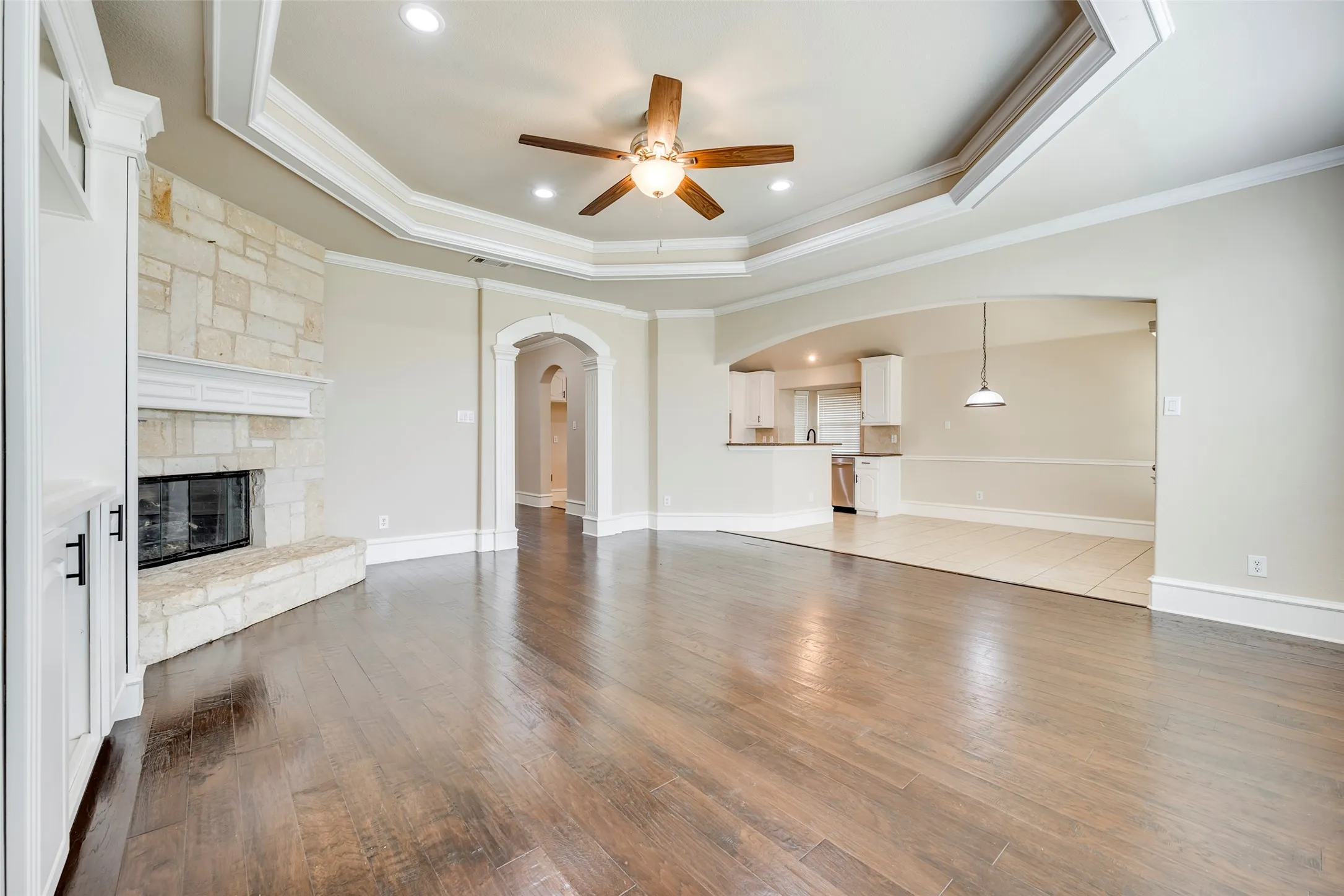Hardwood flooring, tray ceiling with recessed lighting and a ceiling fan, stone fireplace with a mantle, built-in cabinetry with black hardware, and arched doorways