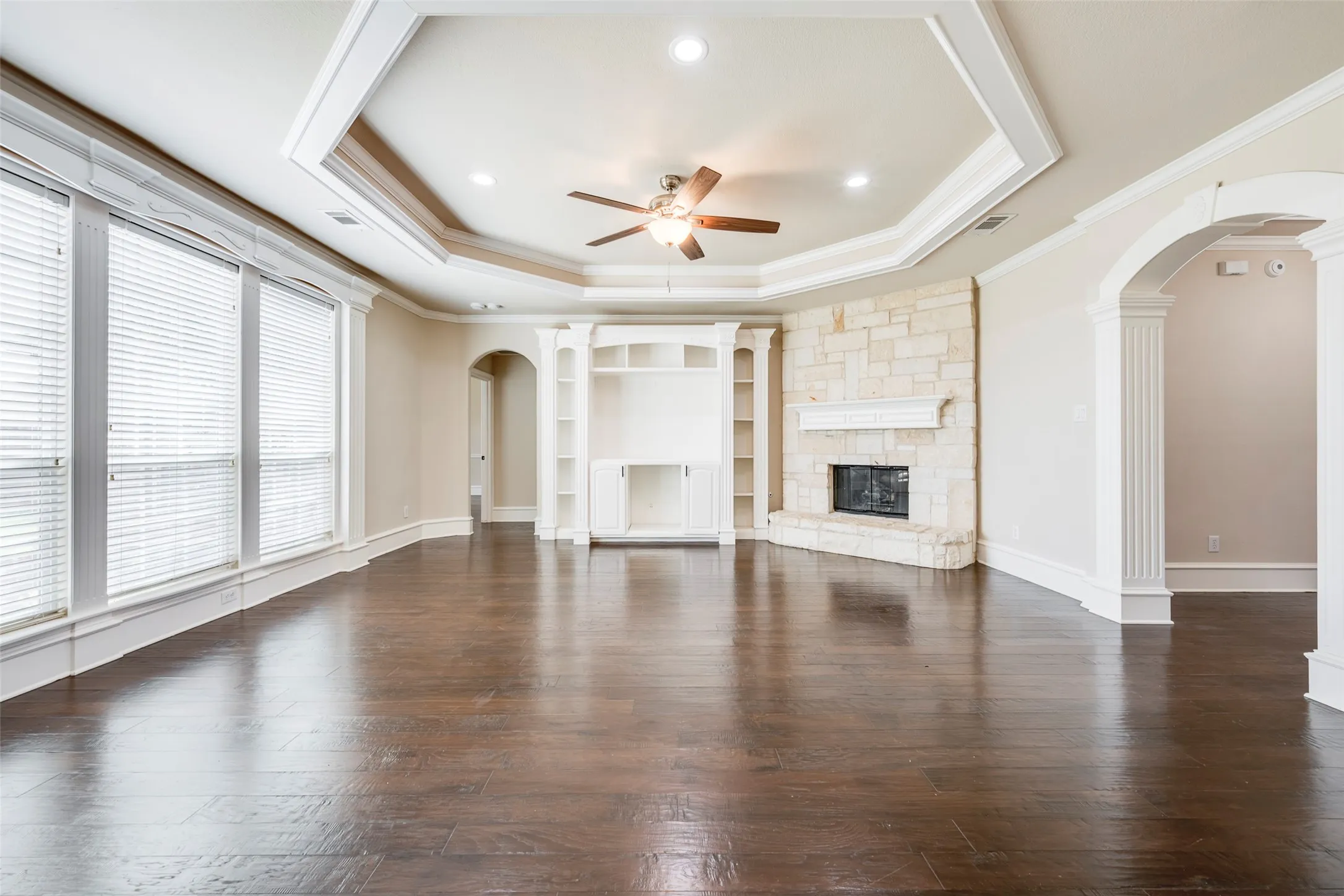 Rich wood-finish flooring, a stone fireplace with a white mantel, built-in shelving, an arched entryway with fluted columns, and a tray ceiling with recessed lighting