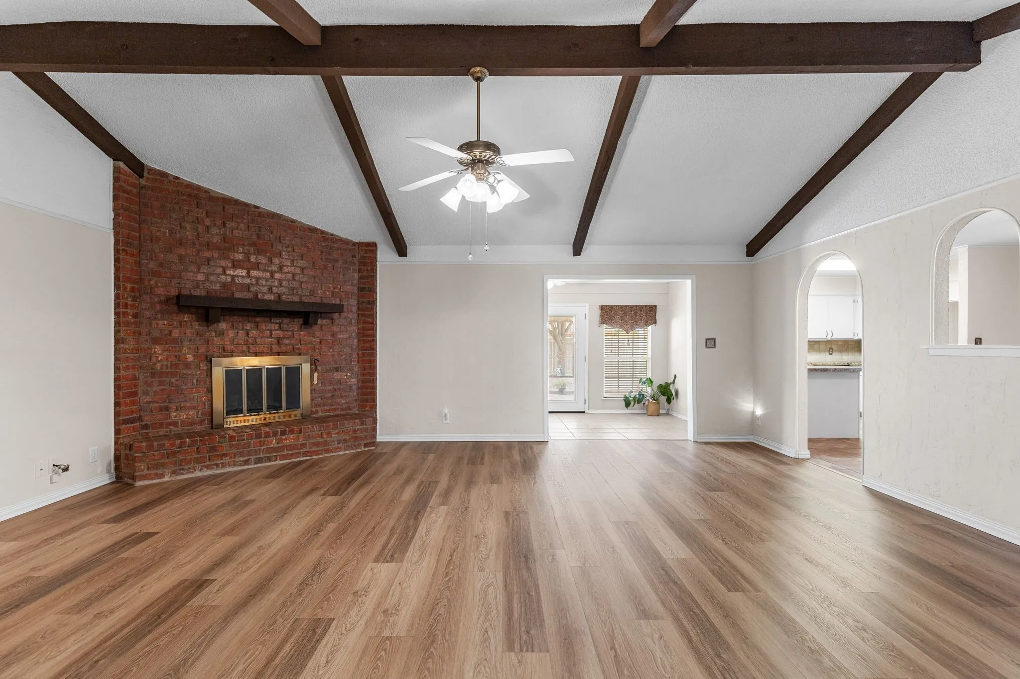 Exposed wood ceiling beams, brick-clad fireplace with brass-finish insert, wood-finish flooring, and arched wall openings