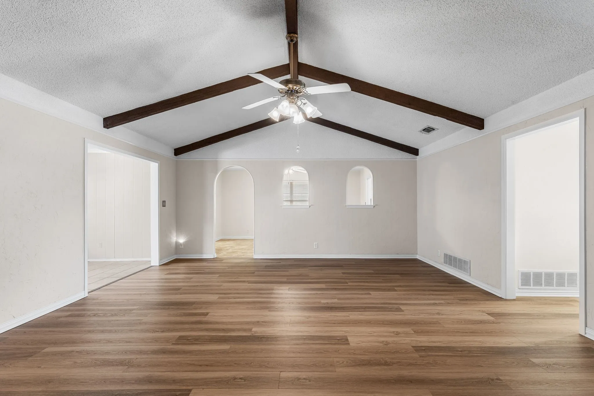 Vaulted ceiling with exposed wood beams, wood-finish flooring, ceiling fan, and arched wall cutouts and walkway to kitchen and dining.