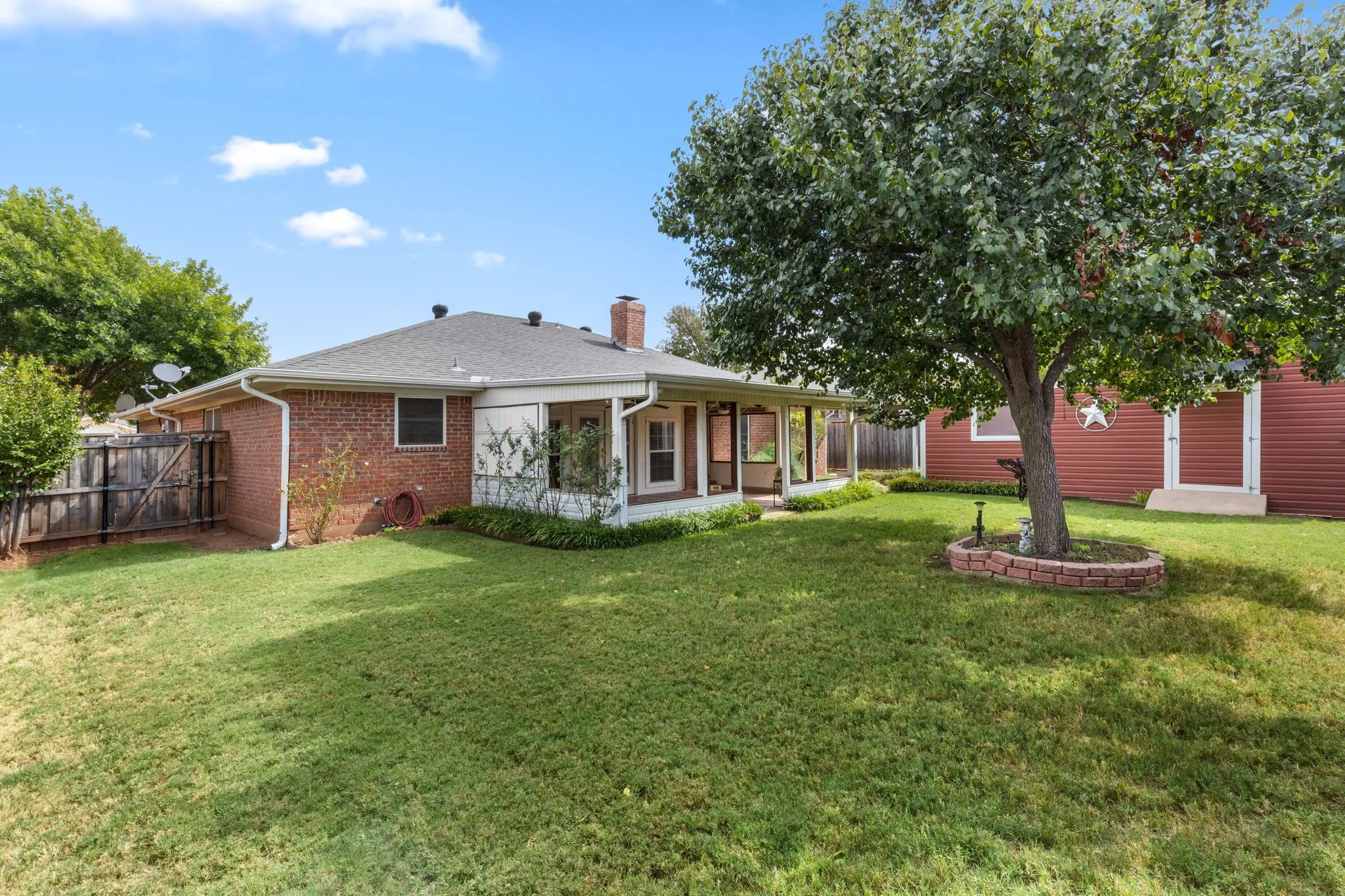 Expansive grassy yard featuring a mature tree with a brick-edged planting bed, a covered patio, and a brick exterior