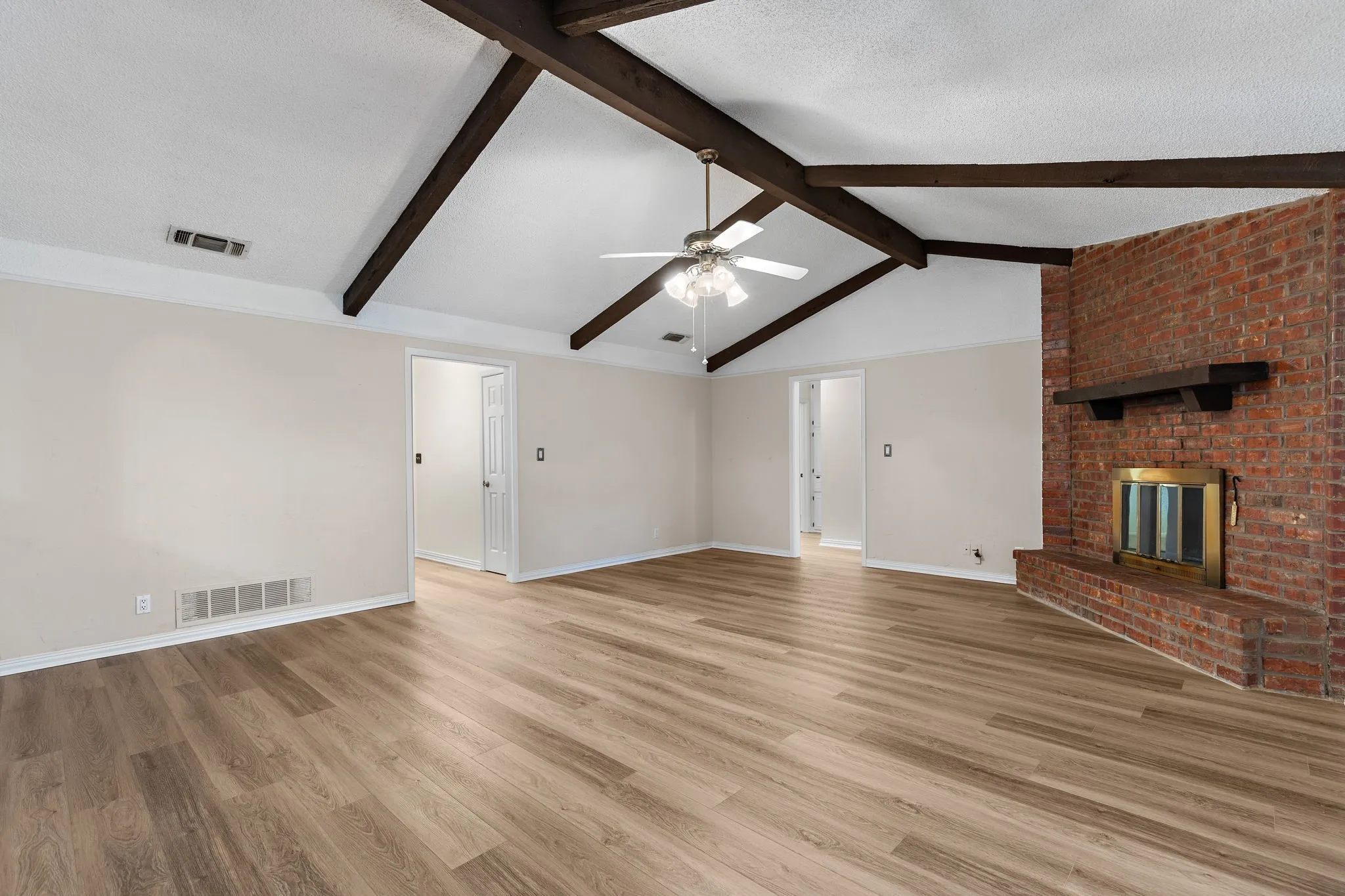 Vaulted ceiling with exposed wood beams, wood-finish flooring, brick-clad fireplace with mantel, and ceiling fan
