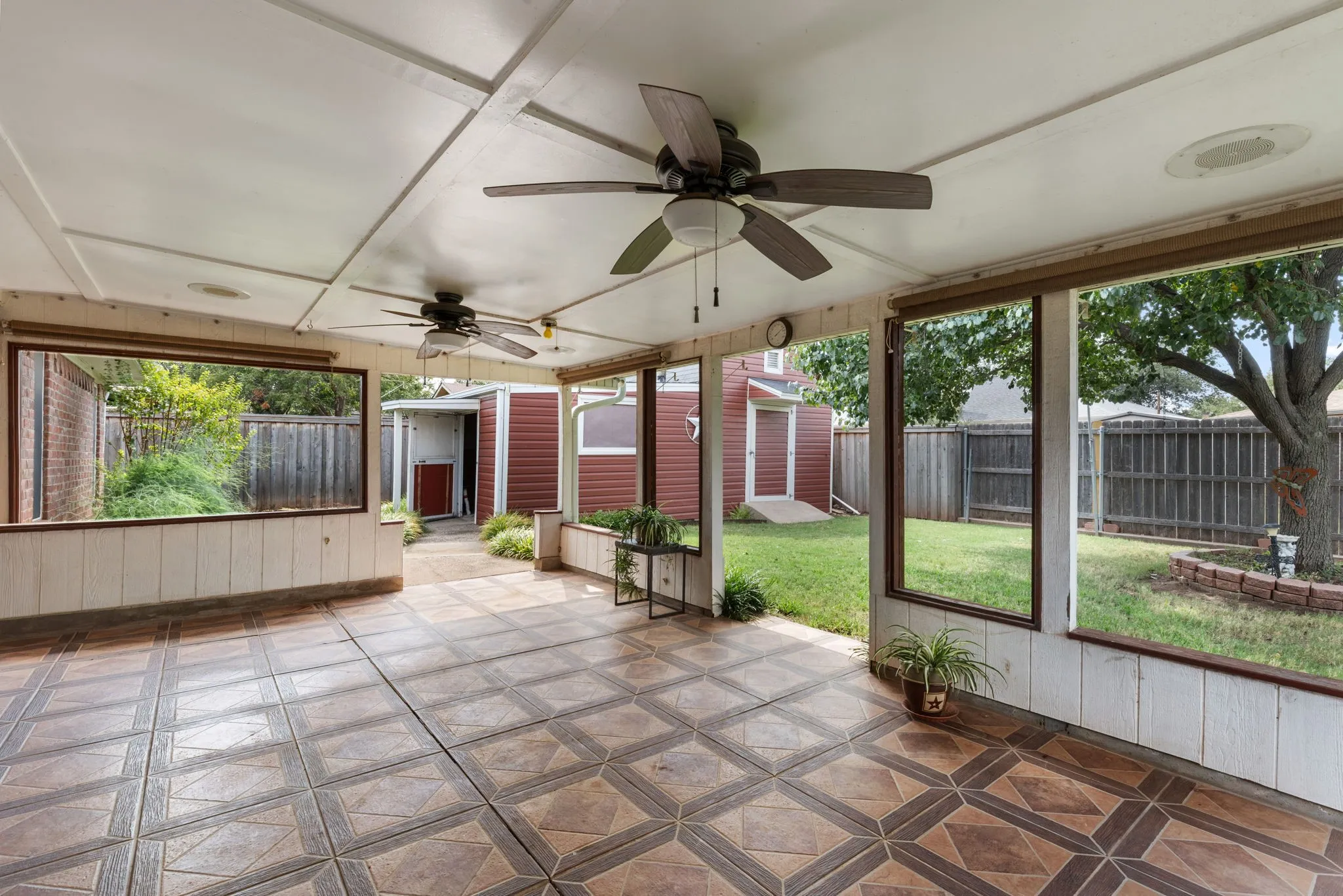 Covered patio featuring patterned tile flooring, wood paneling, multiple ceiling fans, and expansive window cutouts overlooking the backyard. TV wiring and mount on the wall.