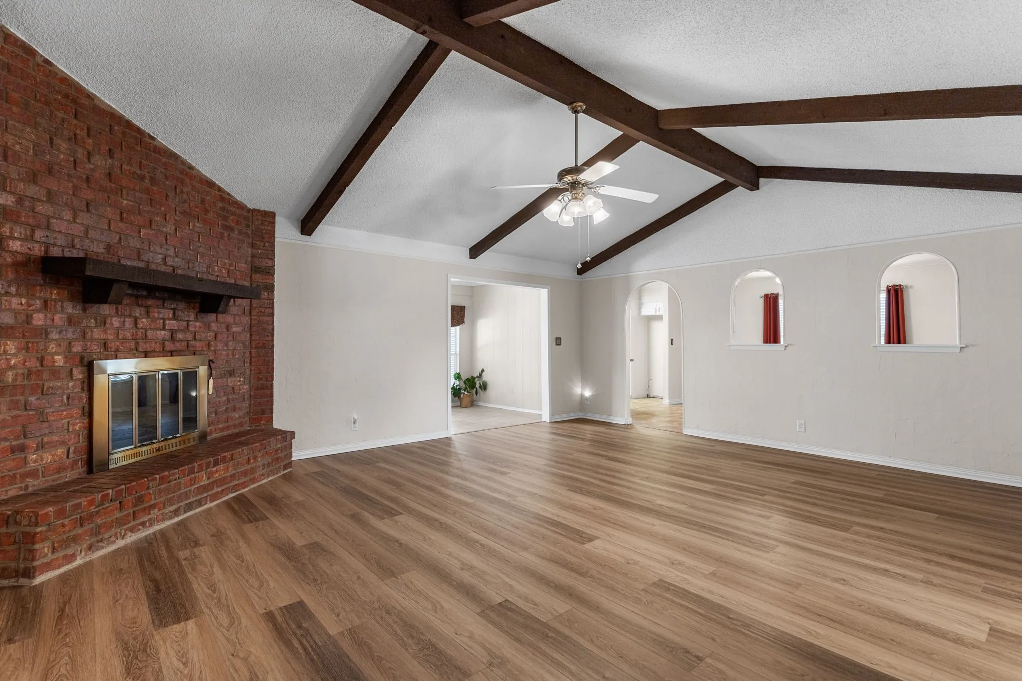 Vaulted ceiling with exposed wood beams, brick fireplace with a wood mantel, wood-finish flooring, and arched wall niches
