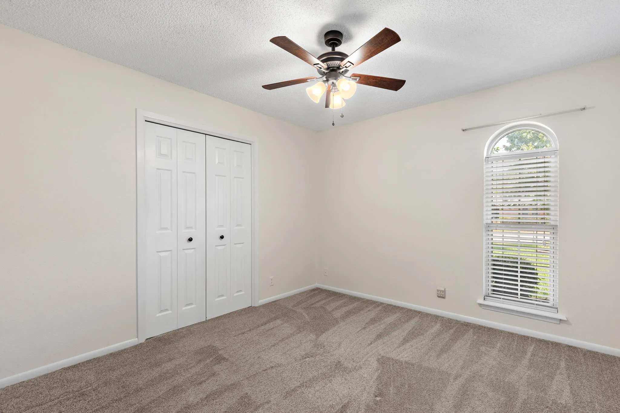 Second Bedroom - Neutral wall tones with carpeted flooring, featuring a ceiling fan with integrated lighting, a bi-fold closet, and a arched window with wood faux blinds