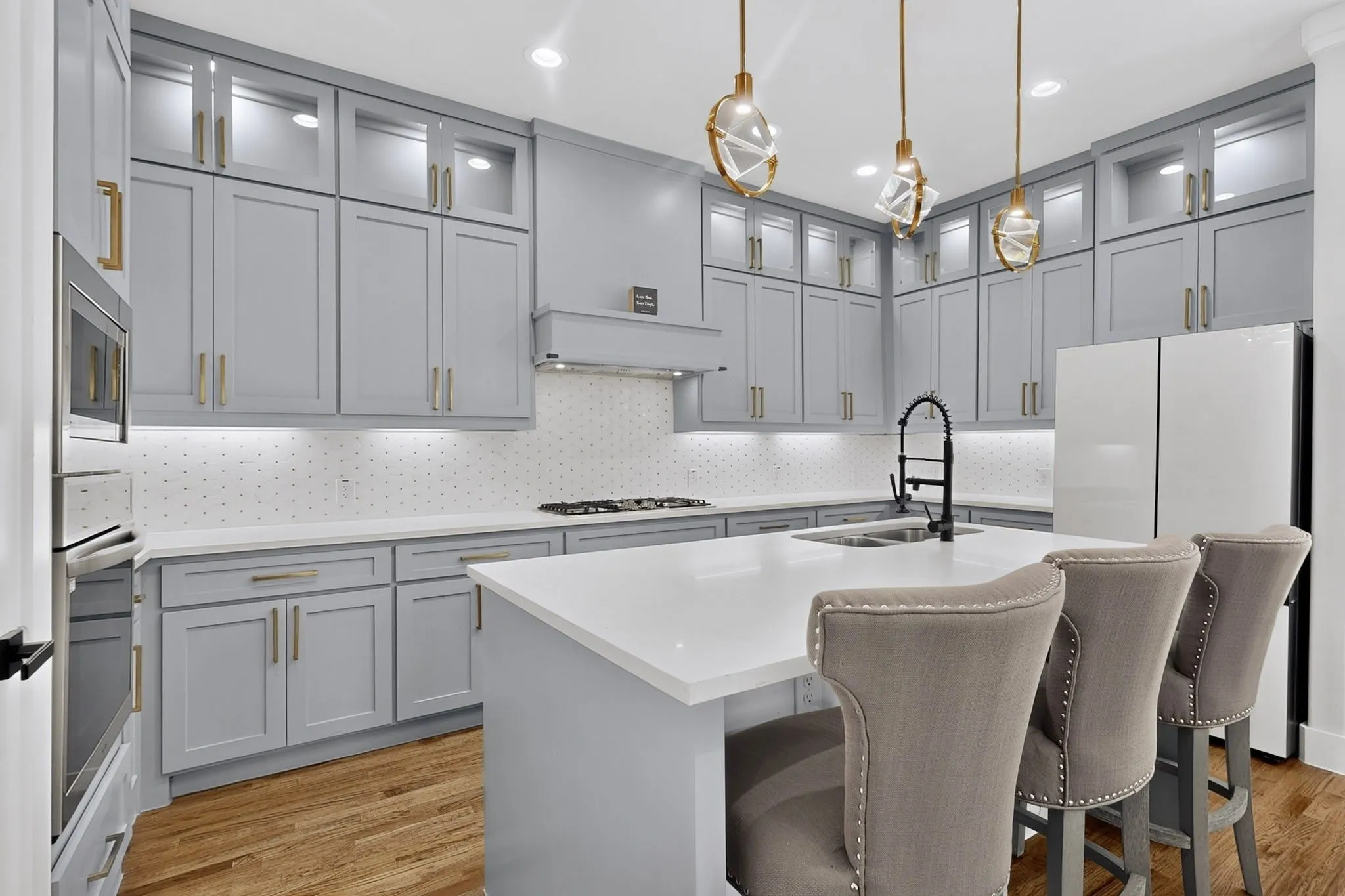 Light gray cabinetry with gold-tone hardware, white countertops, built-in stainless steel appliances, and wood-finish flooring