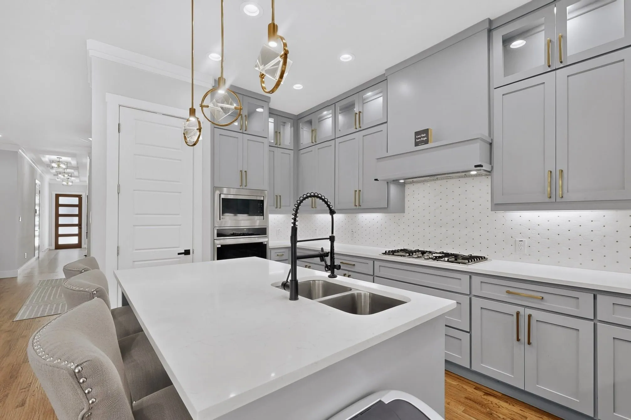 Kitchen featuring a white countertop island with a double sink, light gray cabinetry with gold-tone hardware, a gas cooktop with an overhead range hood, and wood-finish flooring