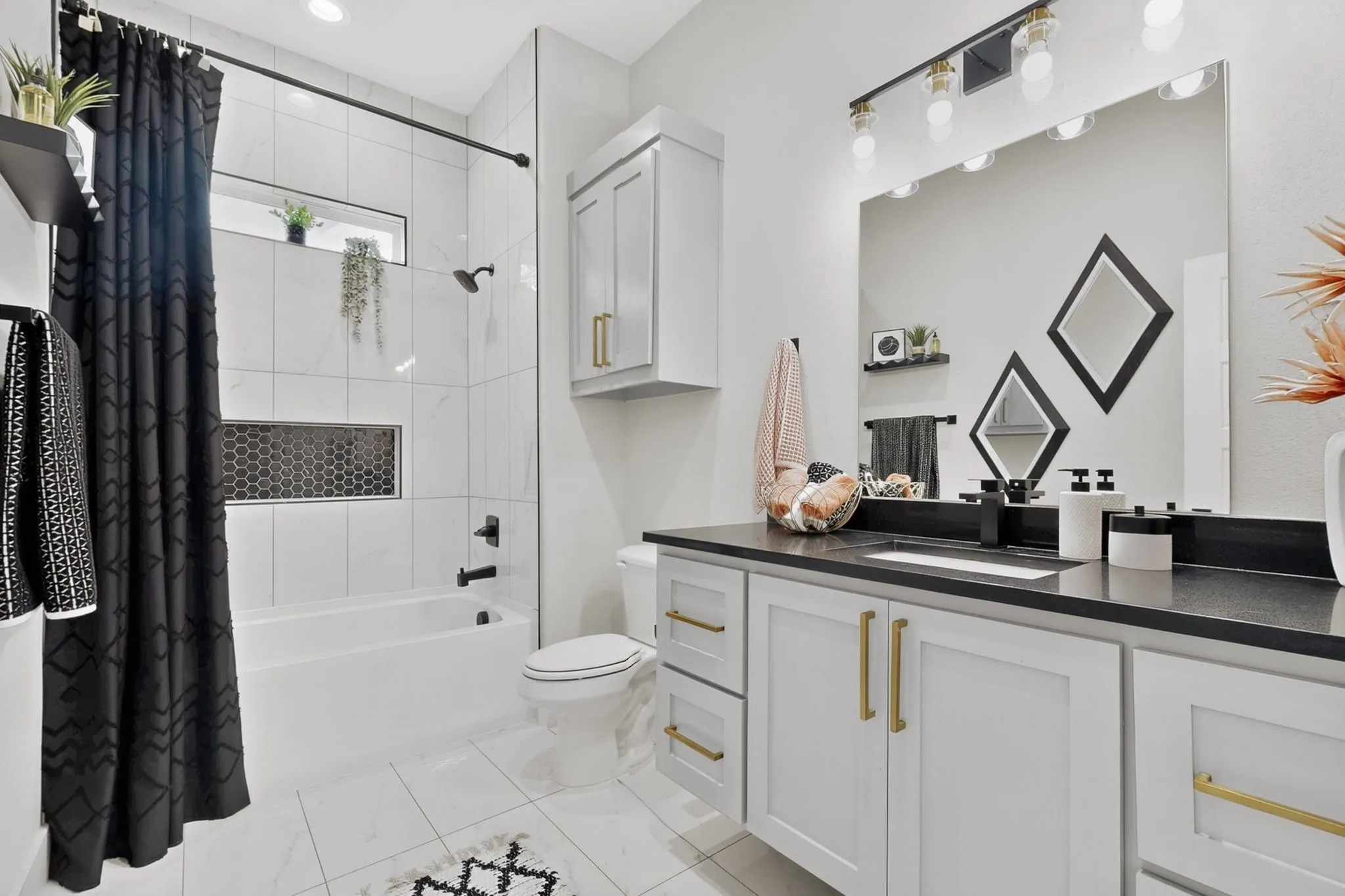 White tile flooring, white vanity with gold-tone hardware, black countertop with undermount sink, built-in bathtub with black fixtures, and a black hexagon tile niche