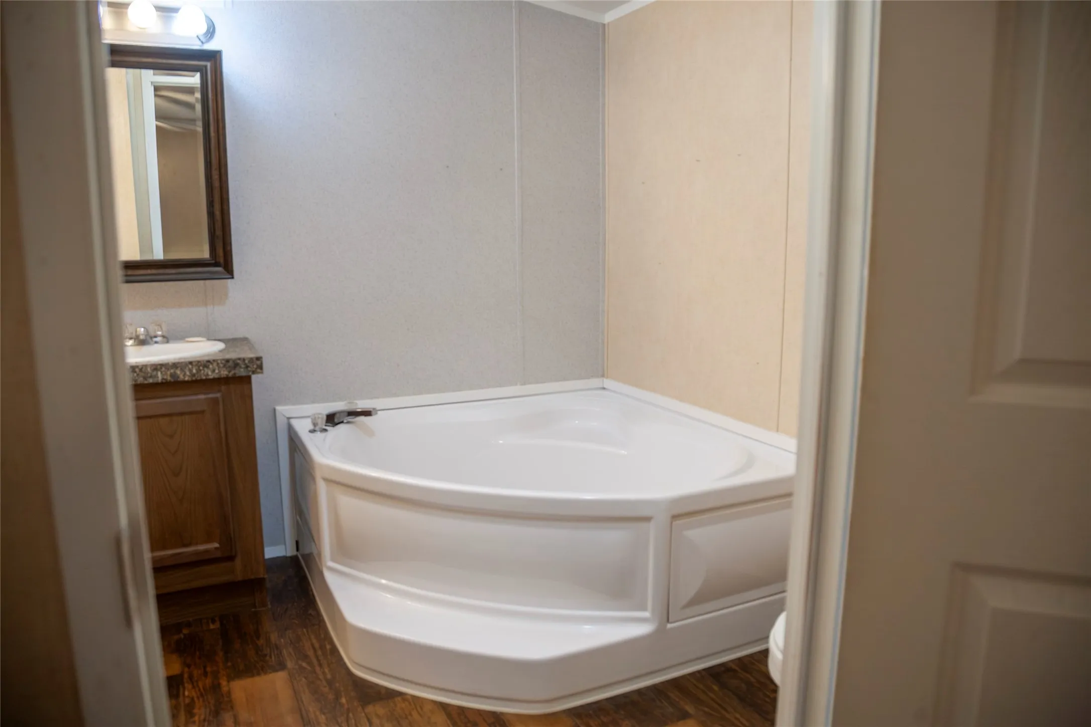 Corner soaking tub with integrated steps, wood-finish flooring, vanity with wood cabinetry, speckled countertop, and a framed mirror