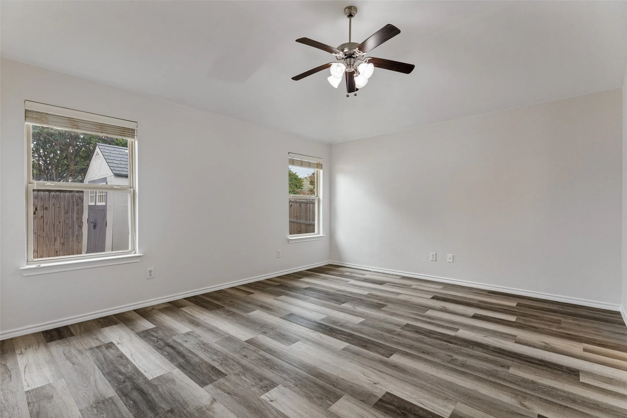 Spacious room with wood-finish flooring, white walls, two windows, and a ceiling fan with integrated lighting