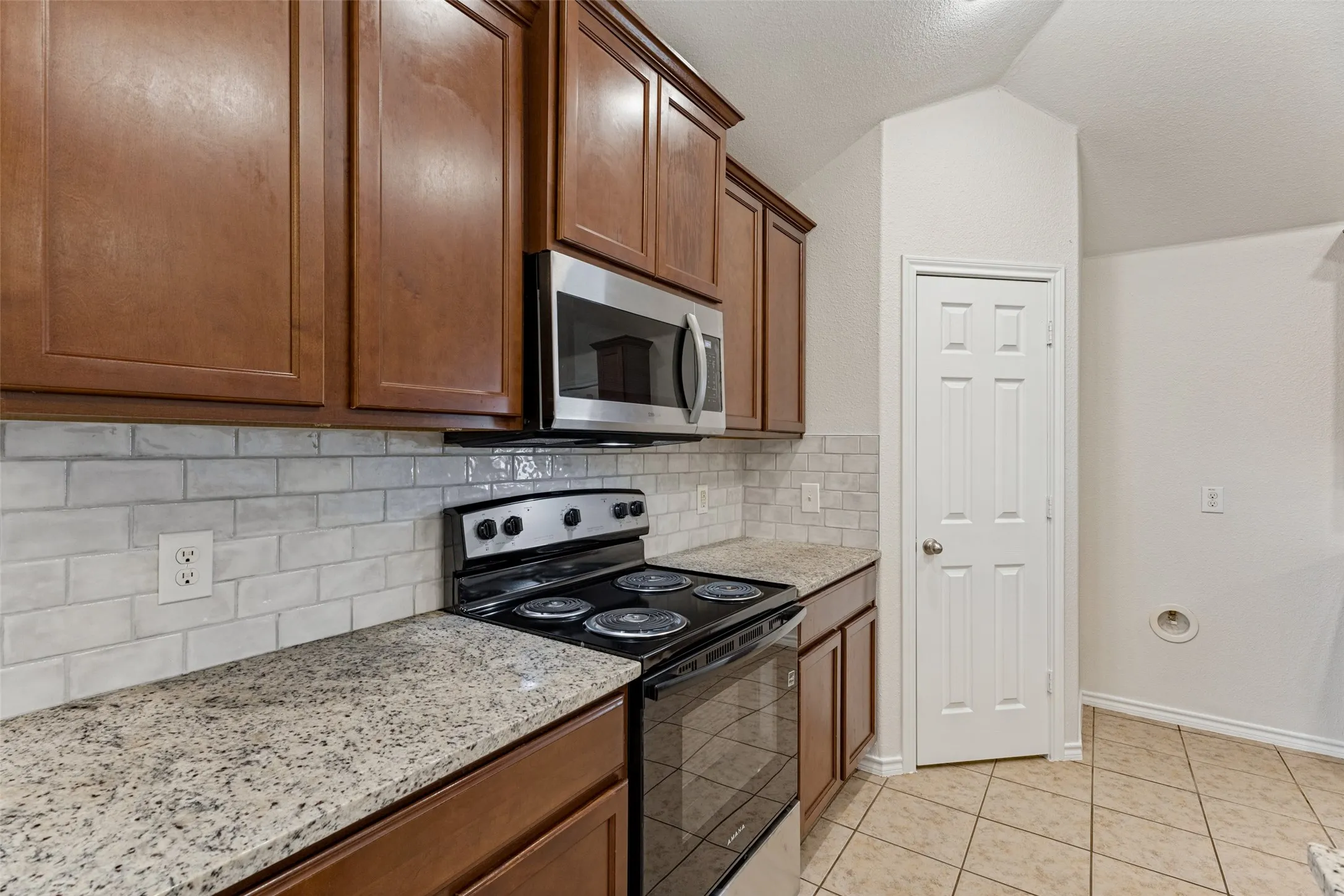 Brown wood cabinetry, subway tile backsplash, granite-style countertops, stainless steel microwave, and tile flooring