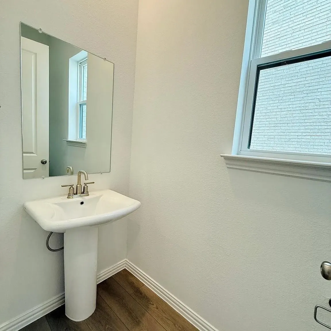 Pedestal sink with a polished faucet, rectangular wall mirror, and wood-finish flooring