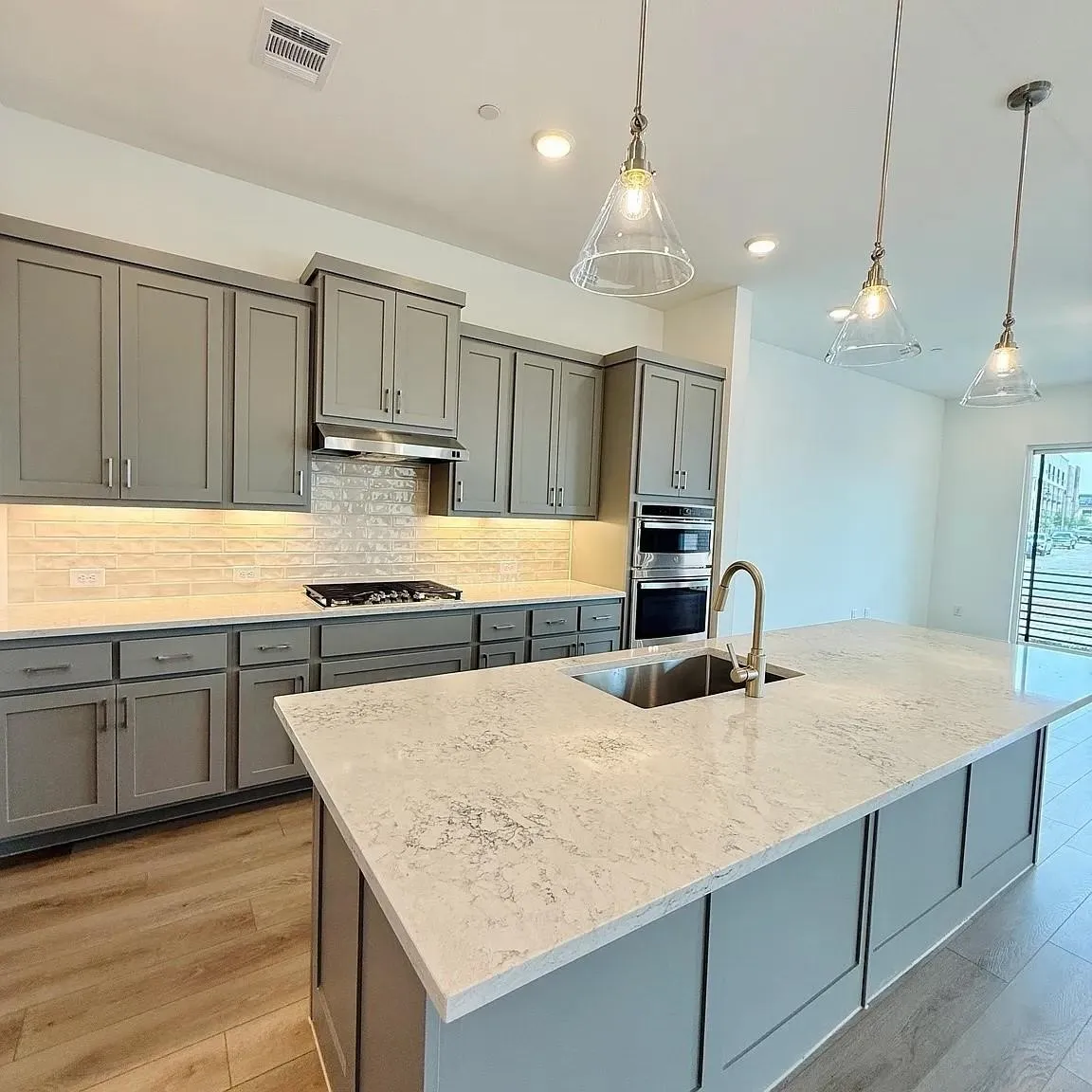 Kitchen featuring gray shaker-style cabinetry, light-toned countertops, a tile backsplash, stainless steel appliances, and a central island with an undermount sink