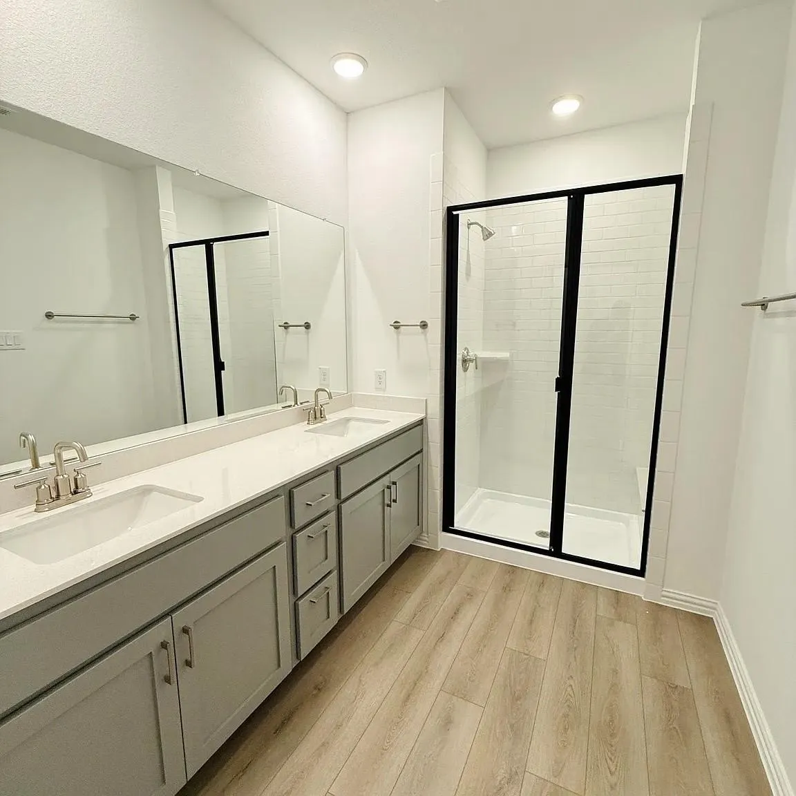 Dual vanity with undermount sinks, light-toned wood-finish flooring, and a walk-in shower featuring a black-framed glass enclosure, white subway tile surround, and a built-in bench