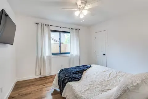 Wood-finish flooring, white painted walls, window with dark trim, ceiling fan with light fixture, and a white door with black hardware