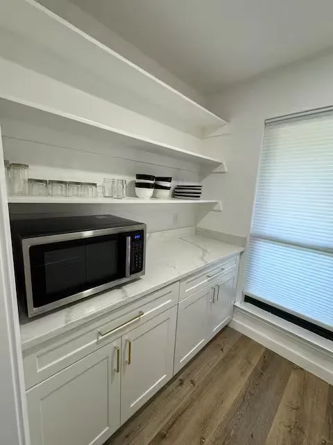 Modern kitchen workspace featuring white cabinetry with brushed nickel hardware, quartz countertops, stainless steel microwave, floating shelves with integrated lighting, and wood-finish flooring