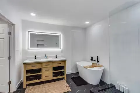Dual vanity with stone countertop and black fixtures, illuminated mirror, freestanding soaking tub, and hexagonal floor tile