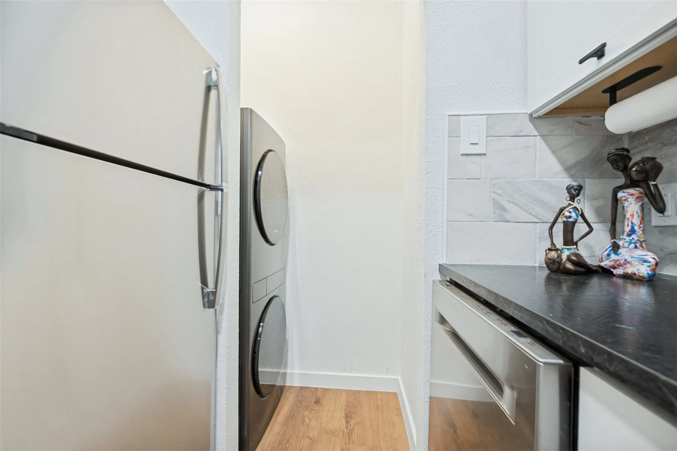 Modern laundry room featuring stacked front-load washers and dryers with sleek metal handles, dark countertop with integrated workspace, white subway tile backsplash, wood-finish flooring, and recessed ceiling lighting