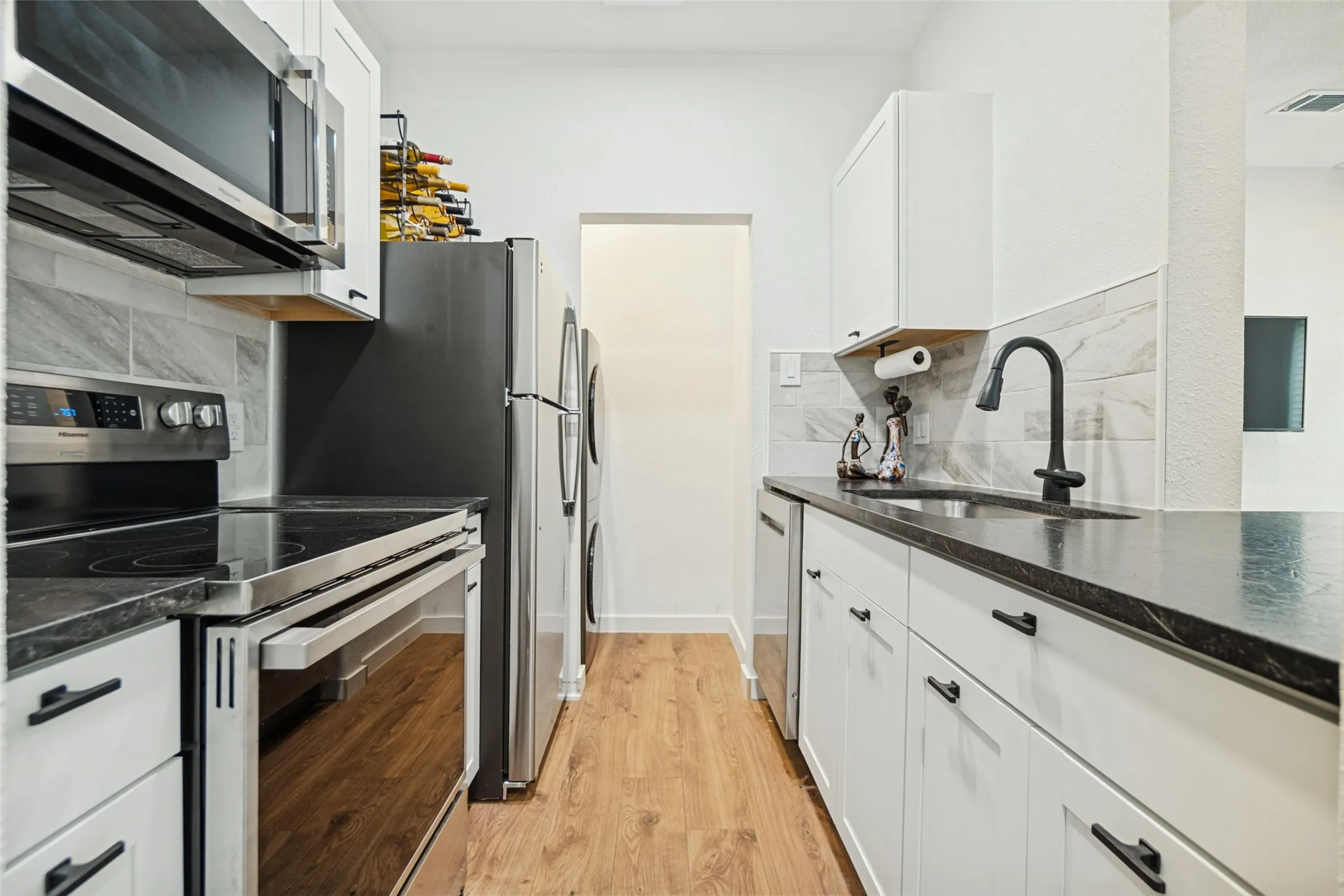 White shaker-style cabinetry, black countertops, wood-finish flooring, stainless steel appliances, and a tile backsplash