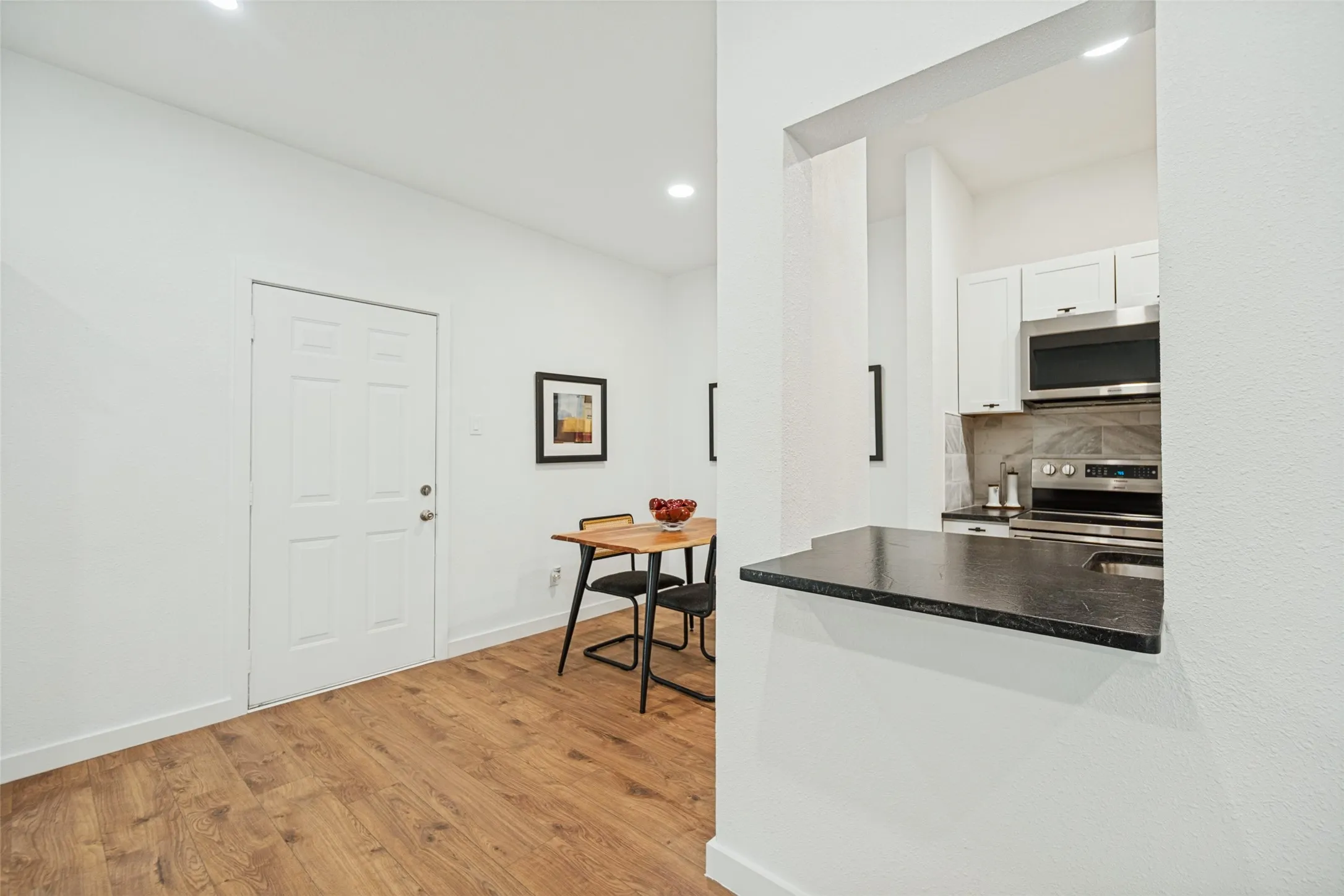 Wood-finish flooring, recessed lighting, white panel door, and a pass-through kitchen window featuring a dark countertop and stainless steel appliances