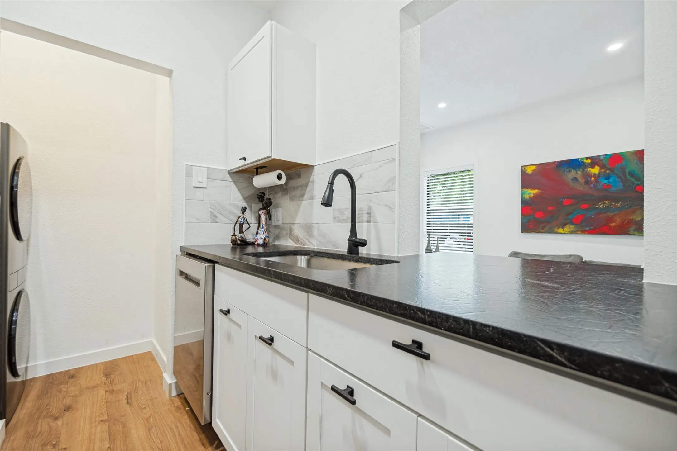 Laundry area featuring wood-finish flooring, stacked front-load machines, white cabinetry with black hardware, dark countertops, and a tile backsplash