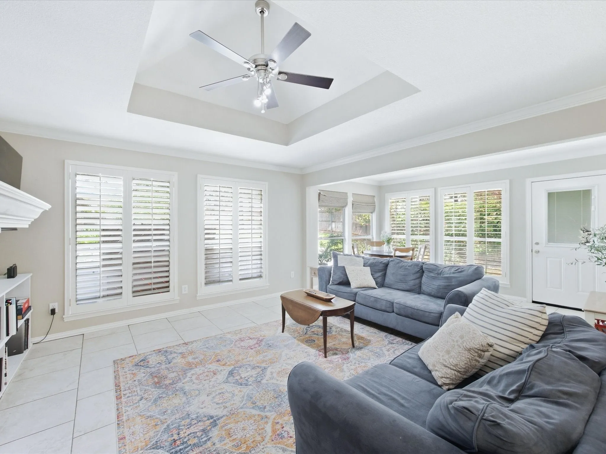 Recessed tray ceiling with ceiling fan, tile flooring, multiple windows with plantation shutters, crown molding, and built-in shelving