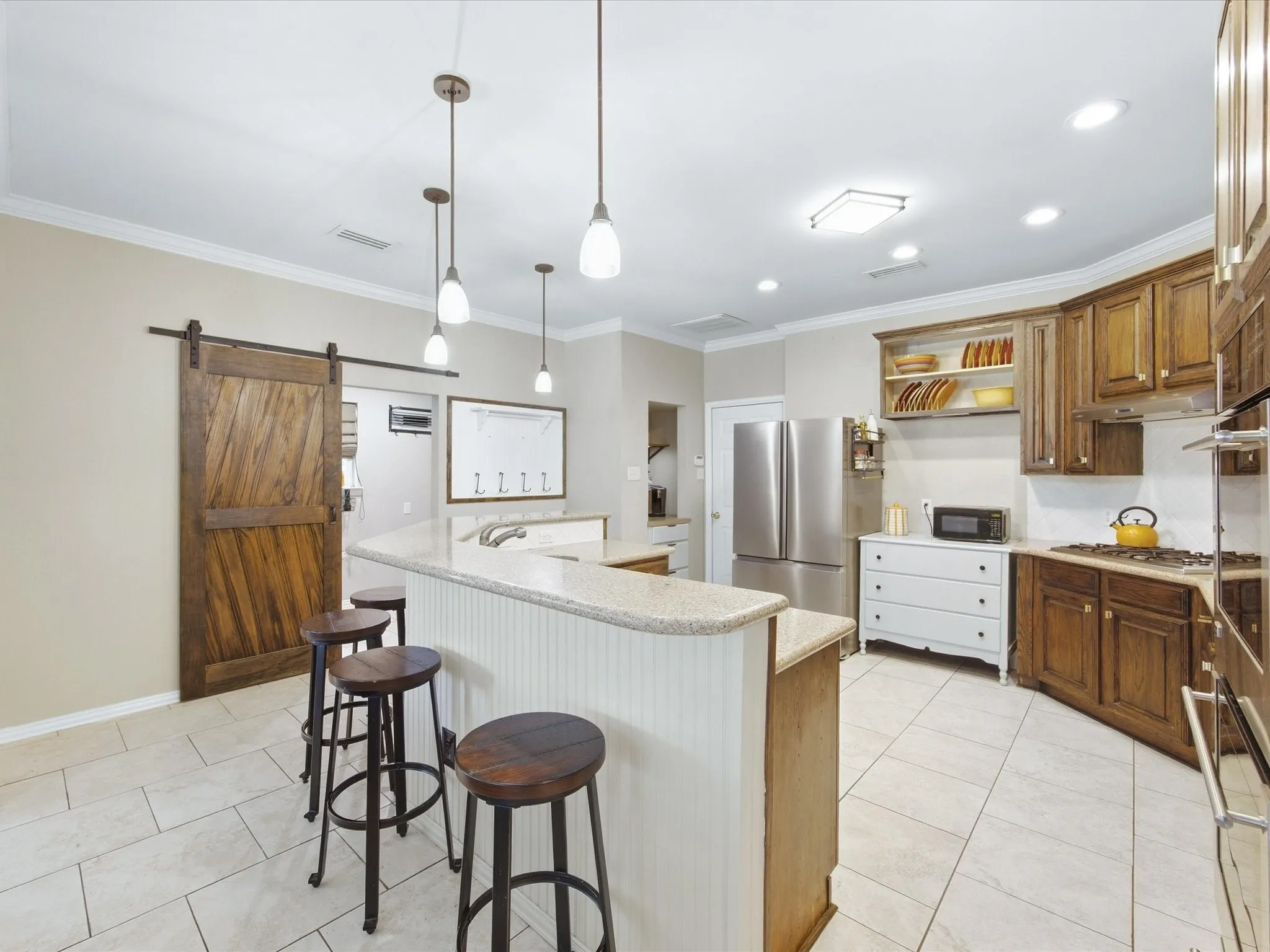 Spacious kitchen featuring a curved island with beadboard paneling, light-colored tile flooring, wood cabinetry, and a sliding barn door