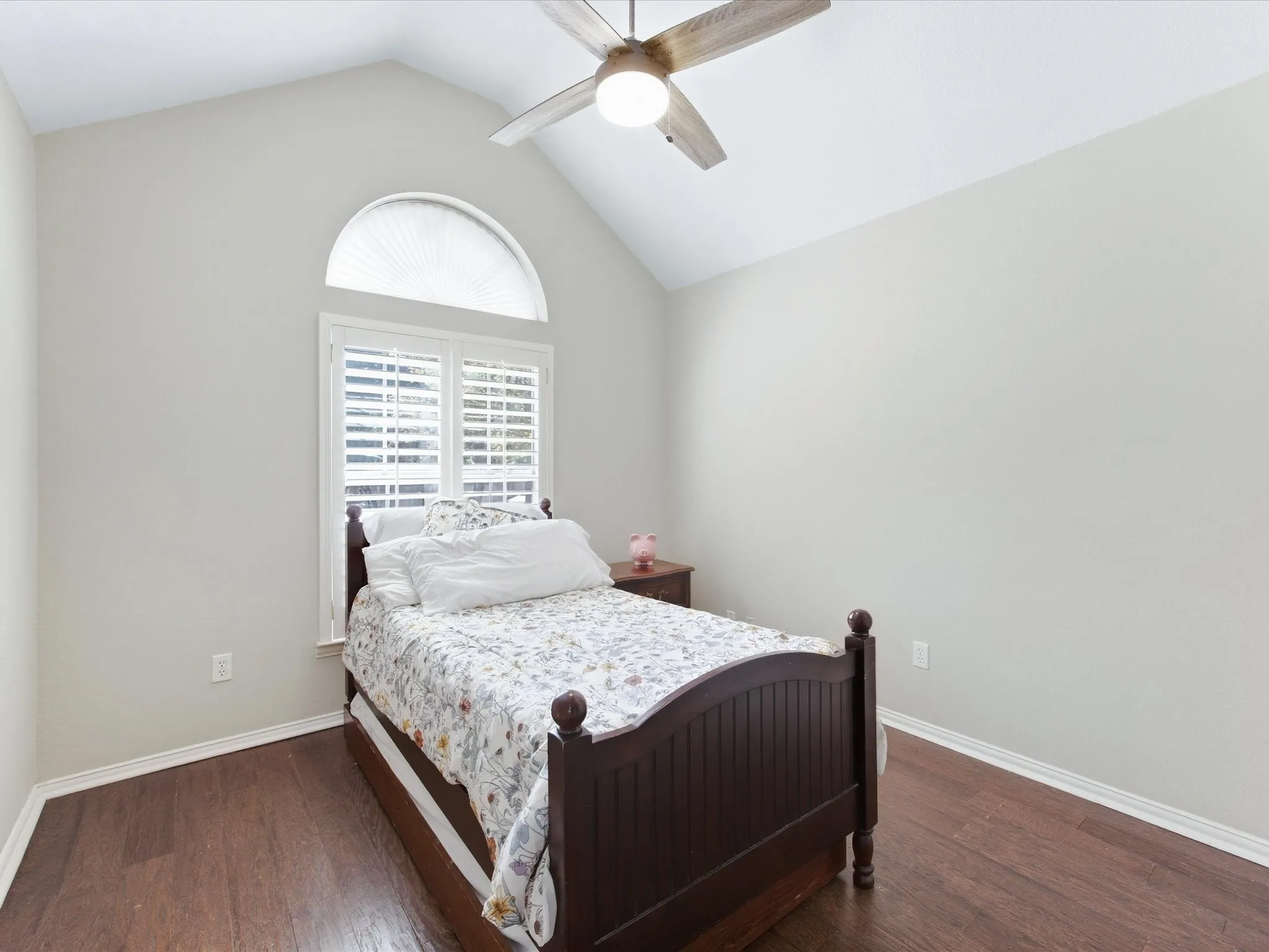 Vaulted ceiling with an installed ceiling fan, an arched window with an installed shade, a rectangular window with installed plantation shutters, and wood-finish flooring