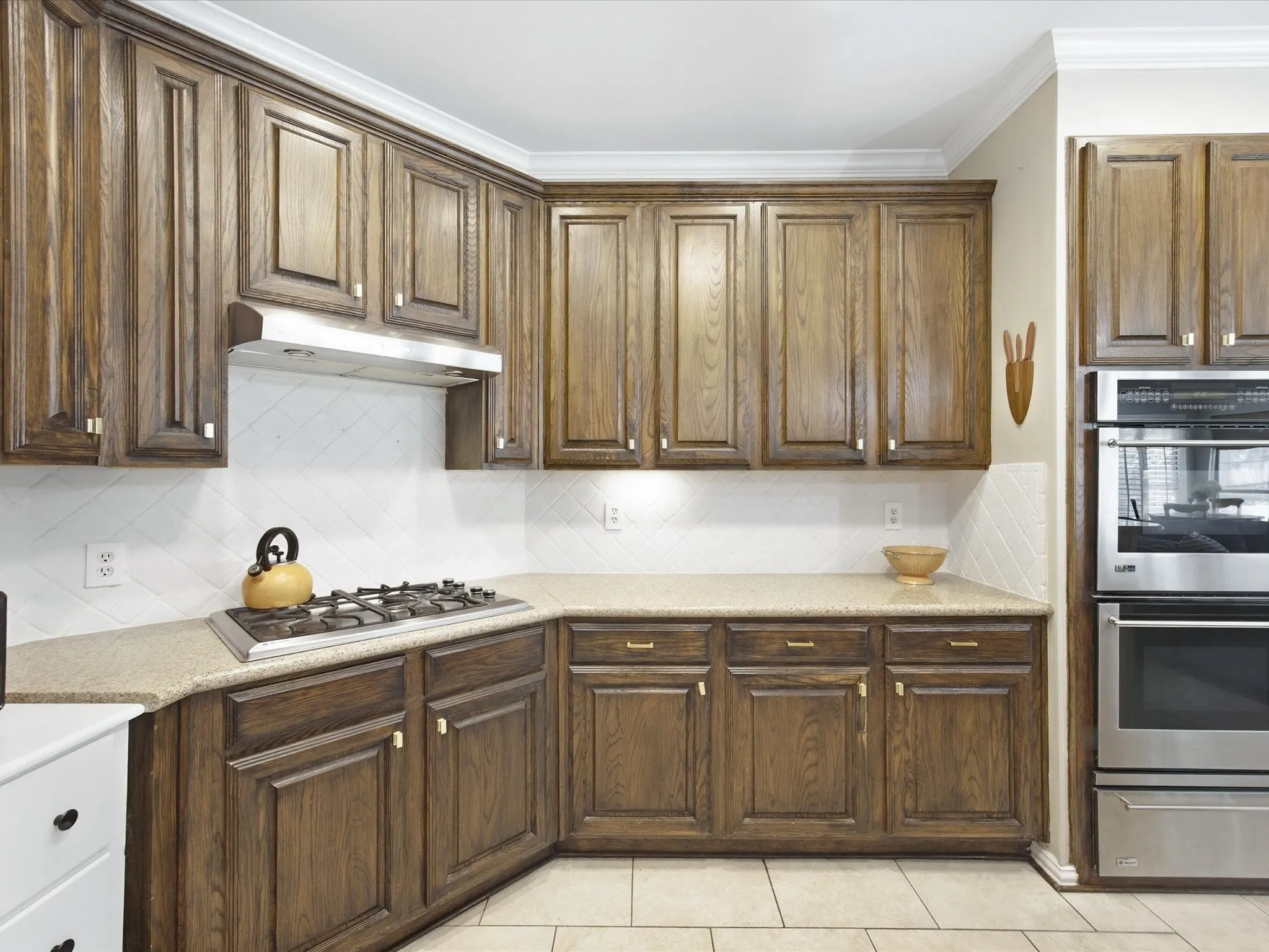 Wood-finish cabinetry, built-in stainless steel double ovens, gas cooktop with stainless steel range hood, stone countertops, and tile flooring