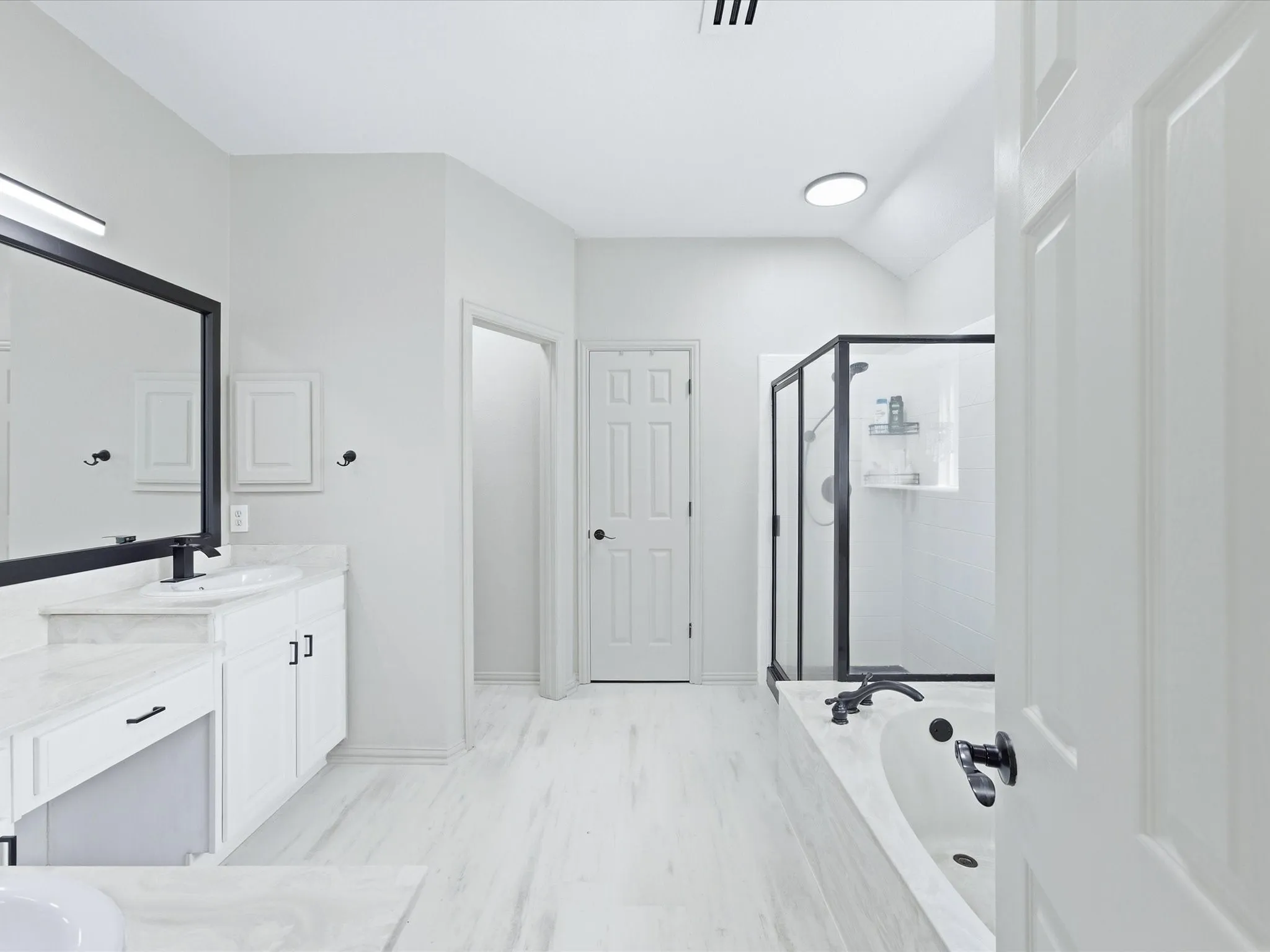 White wood-finish flooring, built-in soaking tub with dark fixtures, walk-in shower with dark frame, dual vanity with white cabinetry, and a circular ceiling fixture