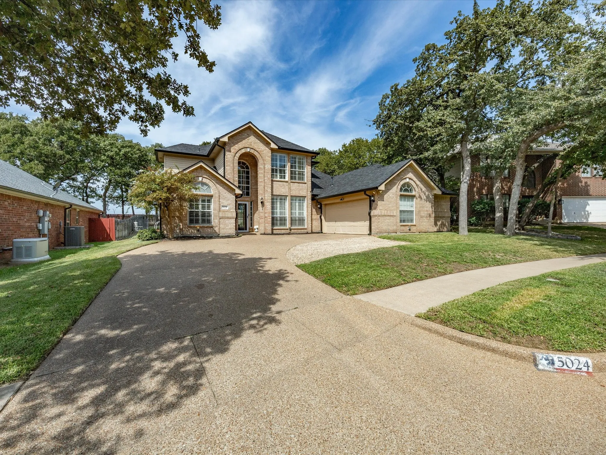 Brick facade with arched window accents, two-story architectural design, expansive paved driveway, attached garage, and mature landscaping