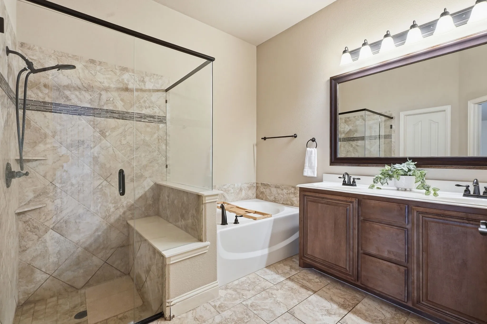 Tiled flooring, dual sink vanity with dark wood-finish cabinetry, built-in bathtub, glass-enclosed shower with a built-in bench, and a rectangular wall mirror