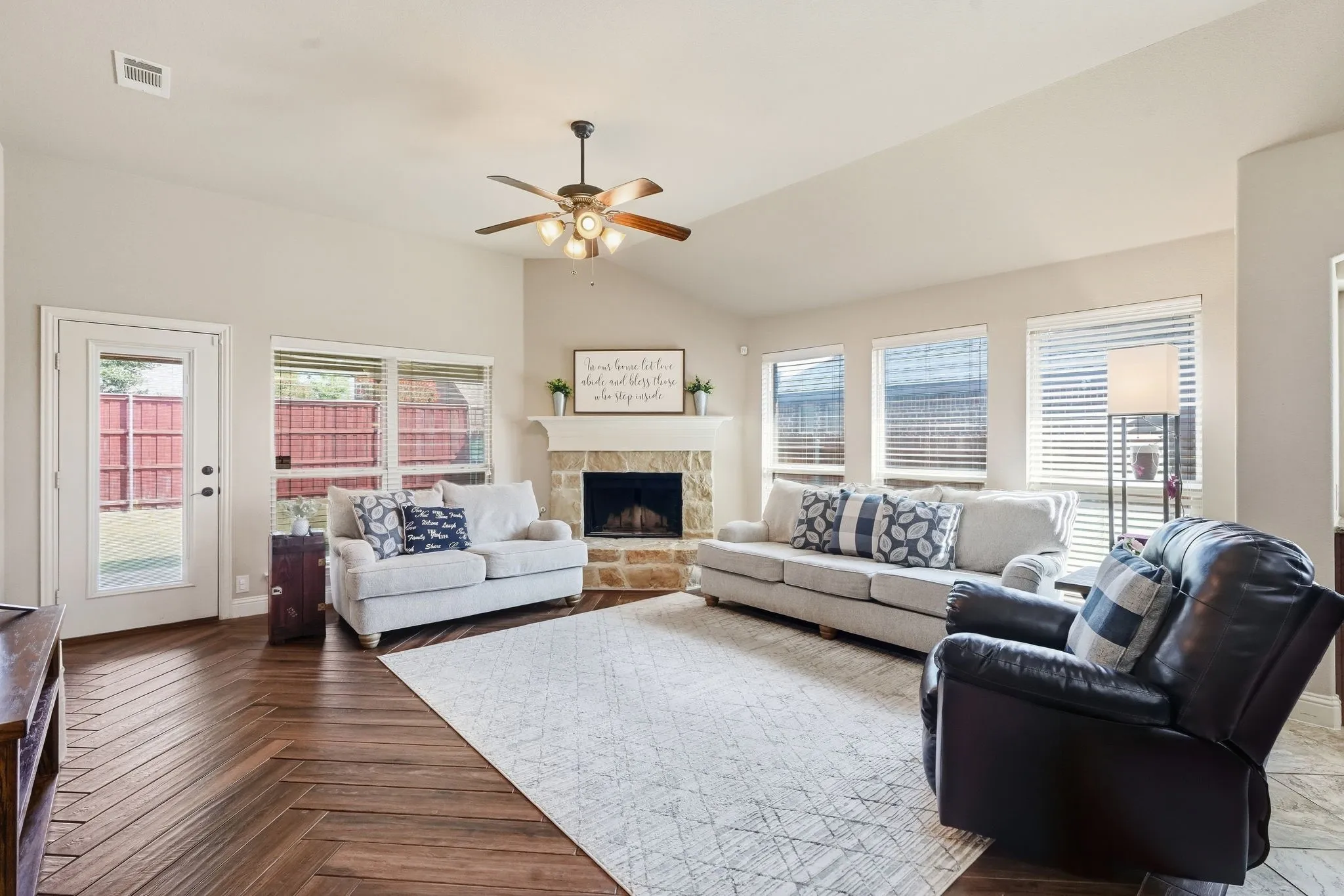 Vaulted ceiling, ceiling fan, stone-clad fireplace, wood-finish flooring, and multiple windows with blinds