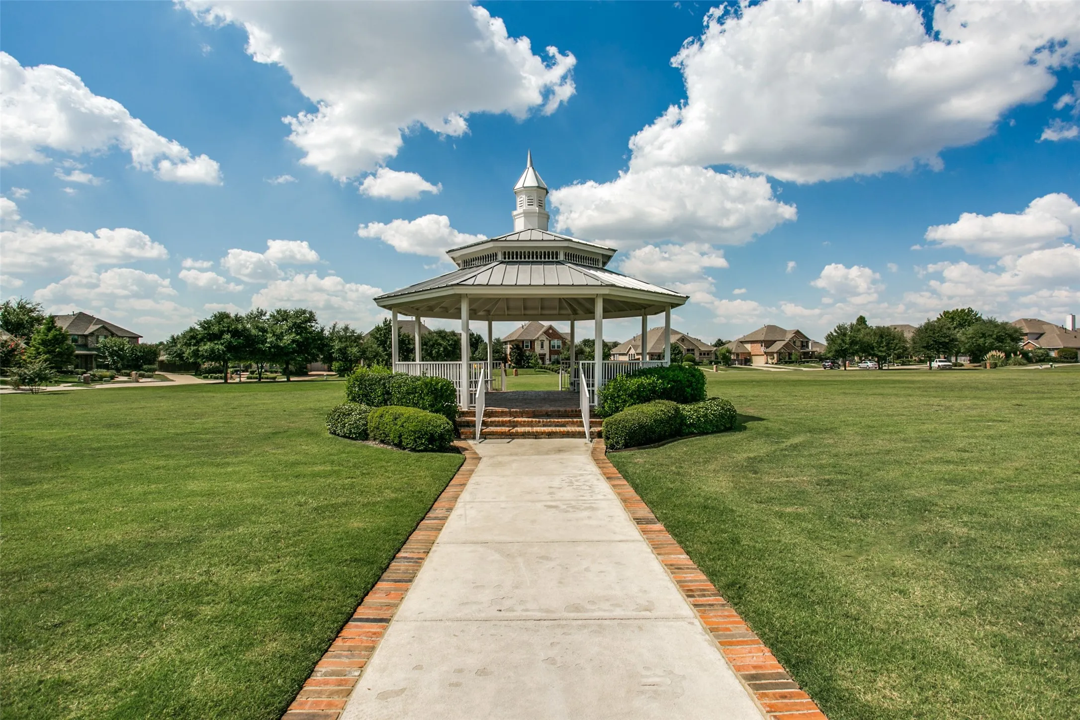 Park just outside the front door has large open green spacw wirh centrally located gazebo with a cupola, surrounded by a manicured lawn, paved walkway, and brick edging
