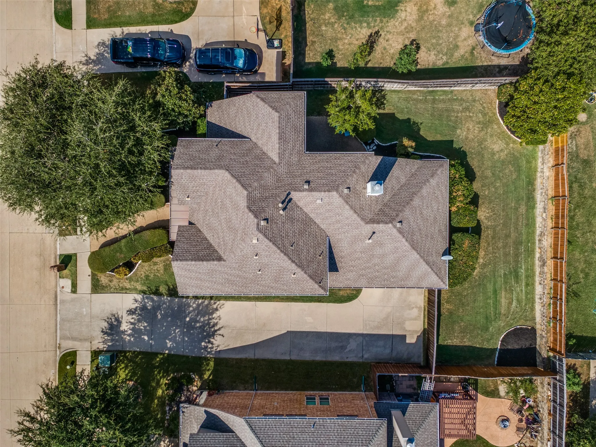 Pitched shingle roof, expansive concrete driveway, mature front yard trees, manicured hedges, and a fenced backyard with grass