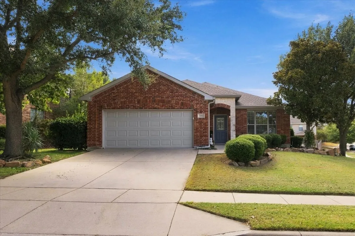 Charming brick home featuring a two-car garage with white panel doors, manicured front lawn with mature landscaping, concrete driveway and walkway, and a gabled roofline with neutral-toned trim