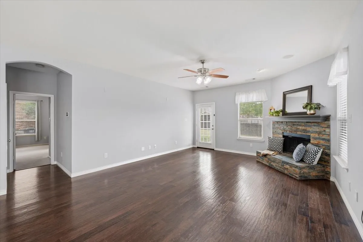 Wood-finish flooring throughout, a stone-clad fireplace with a hearth, a ceiling fan with integrated lighting, and multiple windows