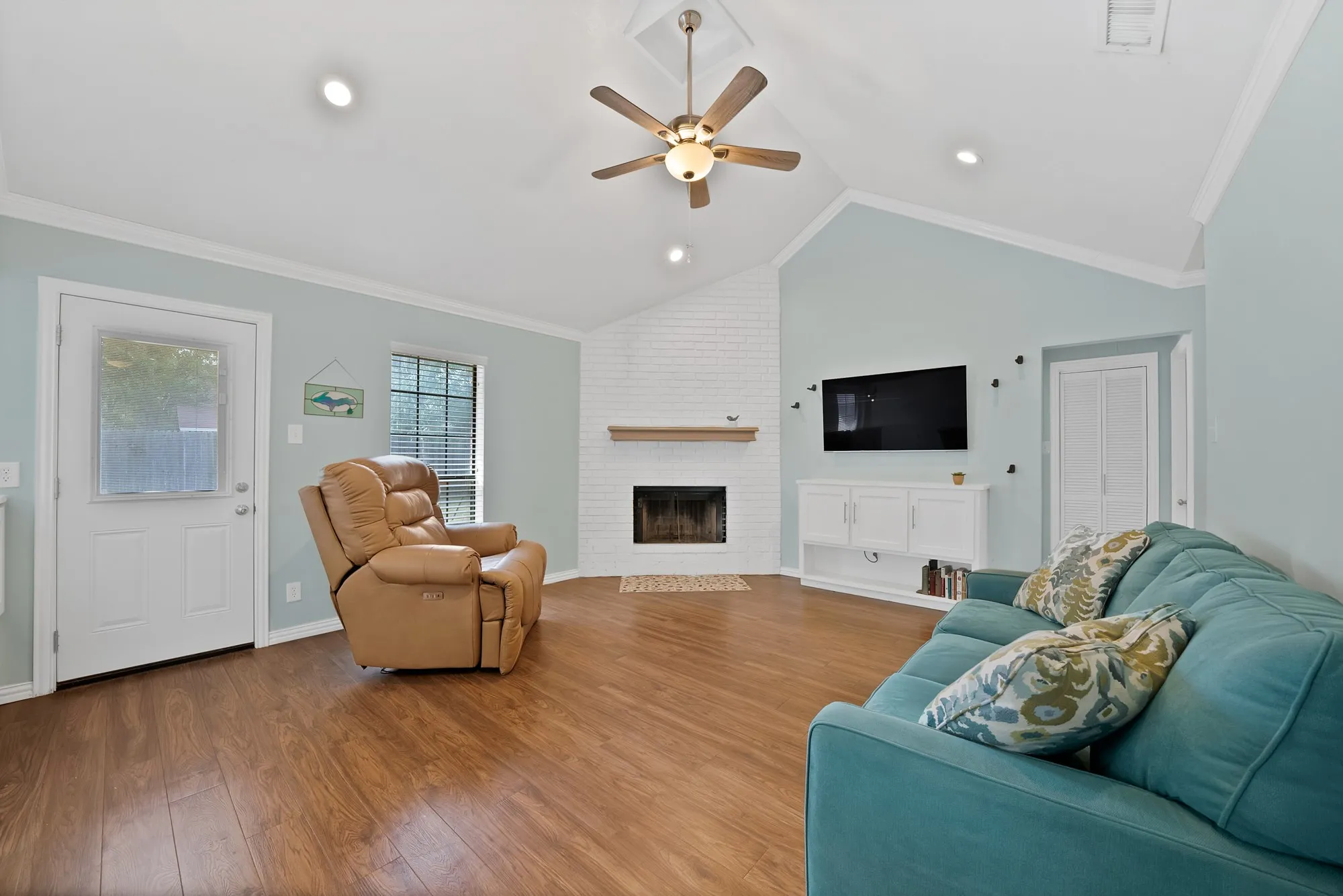 Vaulted ceiling with a ceiling fan, recessed lighting, wood-finish flooring, white brick fireplace with a wood mantel, and built-in white cabinetry