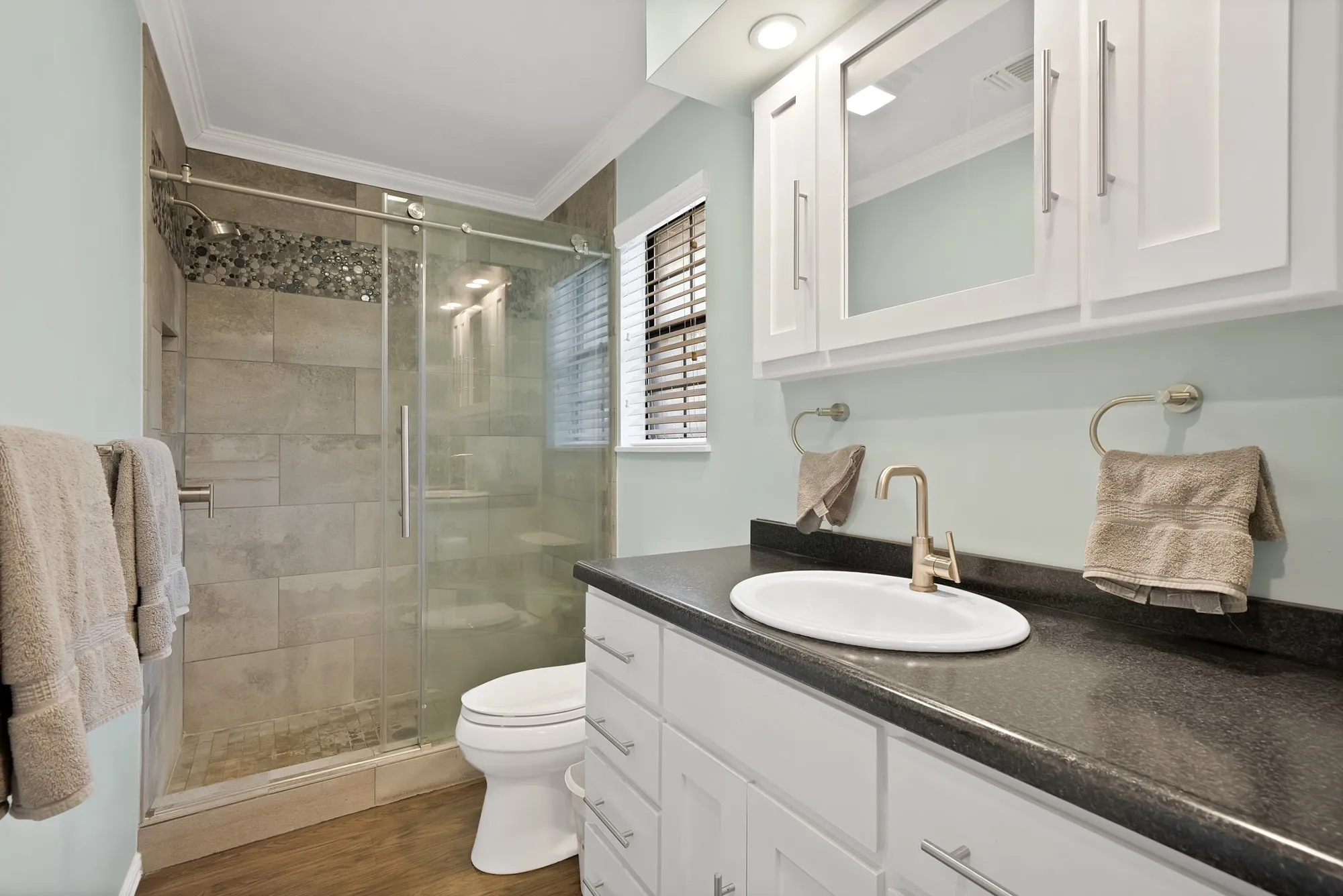 White vanity with a dark countertop and integrated sink, brushed nickel faucet, glass-enclosed shower with tile surround, and wood-finish flooring