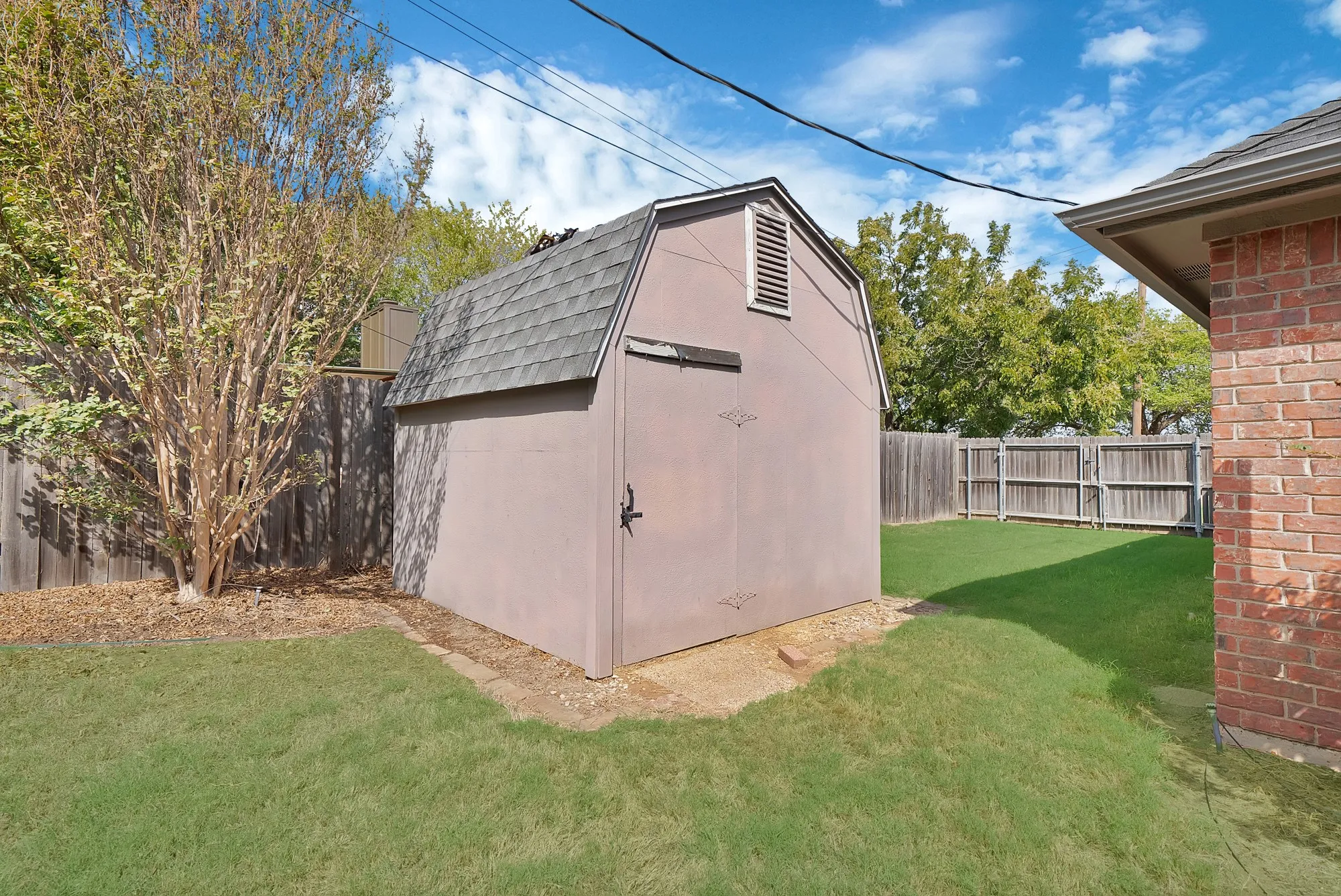Detached storage shed with gambrel roof, brick exterior wall, and wood privacy fencing
