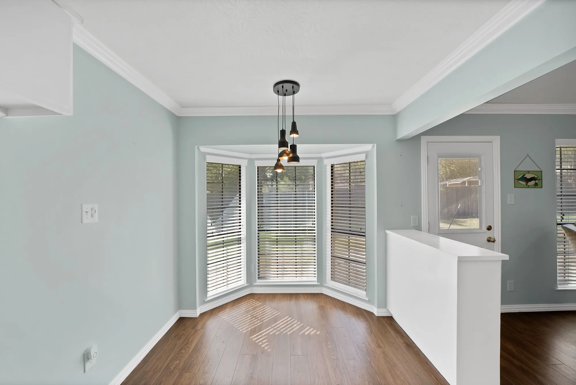 Bay window with blinds, wood-finish flooring, contemporary pendant lighting, crown molding, and a built-in half wall