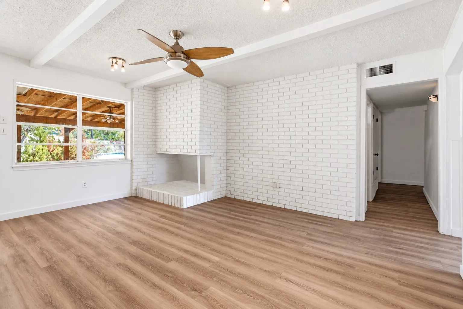 Spacious living area featuring wood-finish flooring, a white brick fireplace, and exposed ceiling beams