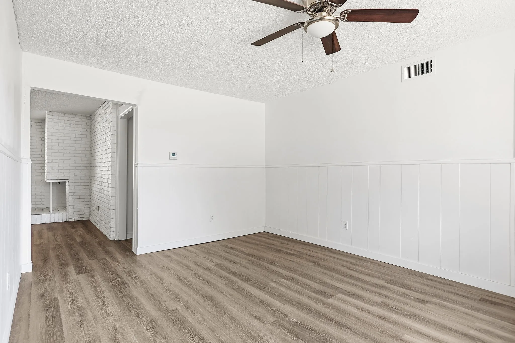 Wood-finish flooring with white wainscoting, ceiling fan, and painted brick fireplace